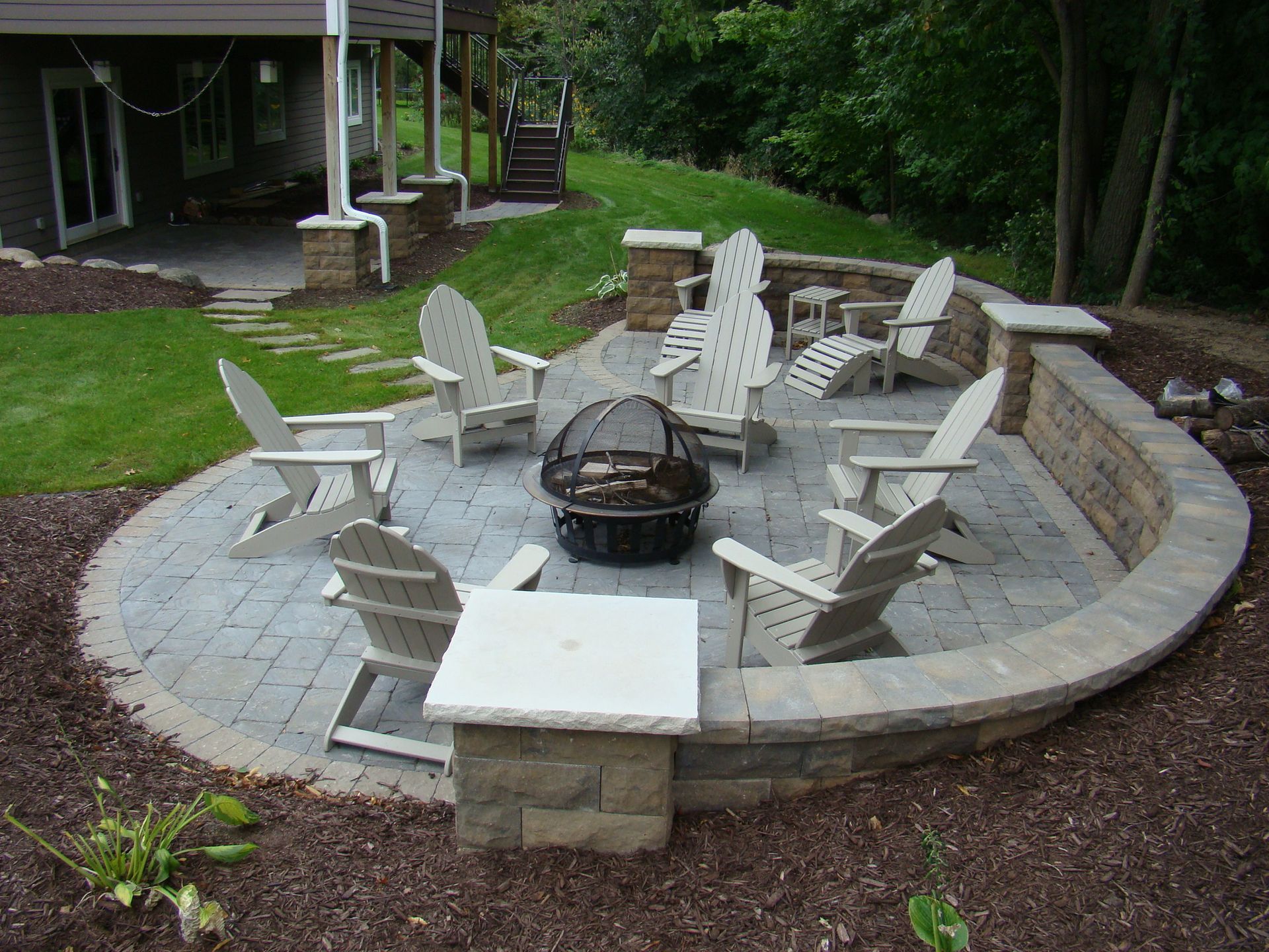 A circular patio with Adirondack chairs around a fire pit, next to a grassy hill and house.