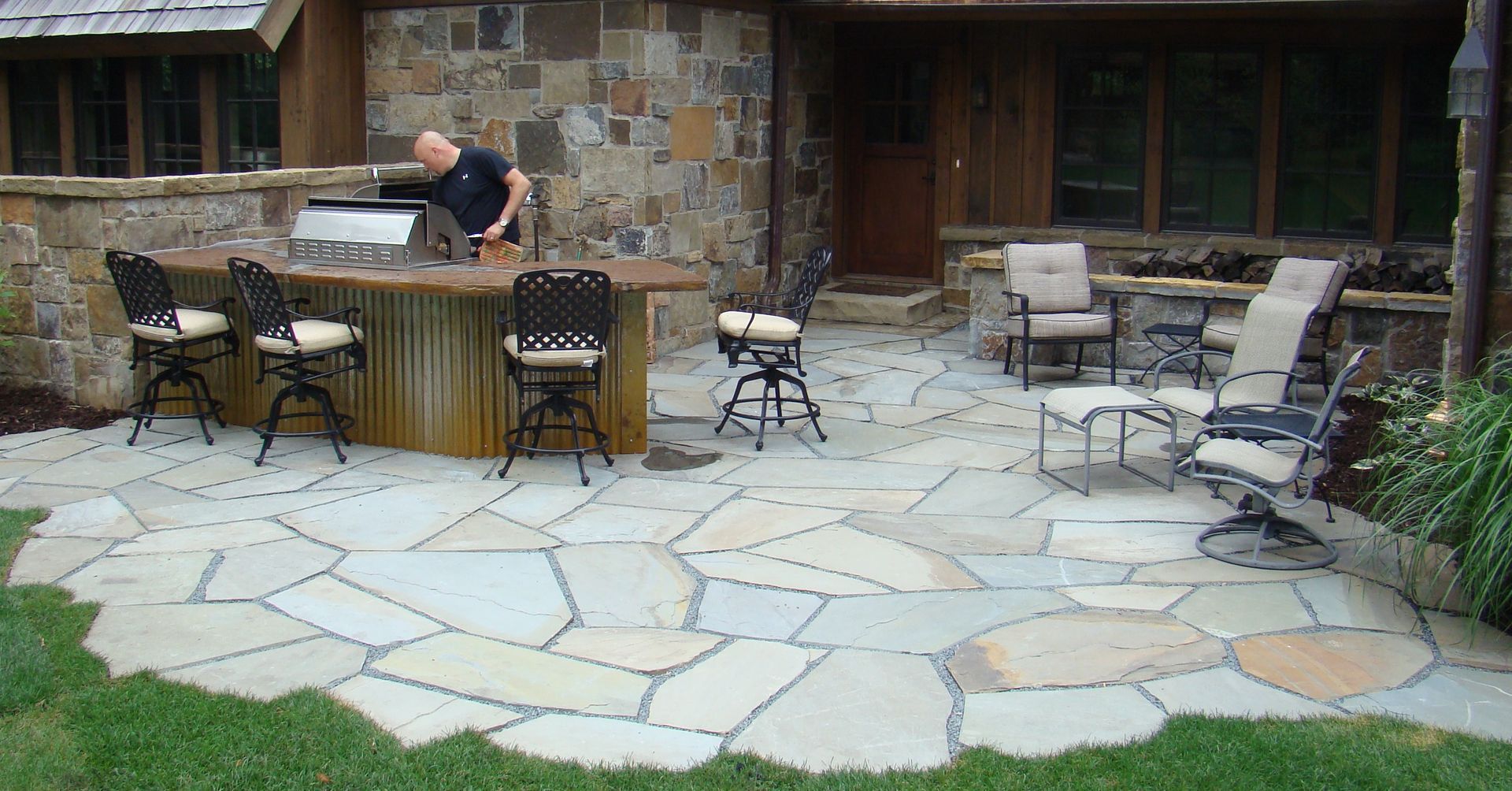 Stone patio with outdoor bar, seating, and a person using the grill near a stone building.