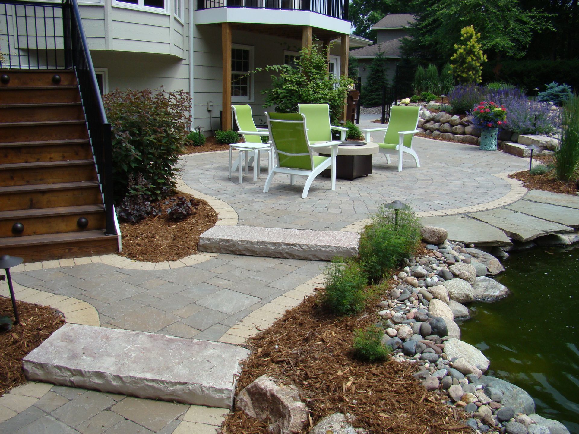 Stone patio with seating area, steps, and pond, surrounded by landscaping.