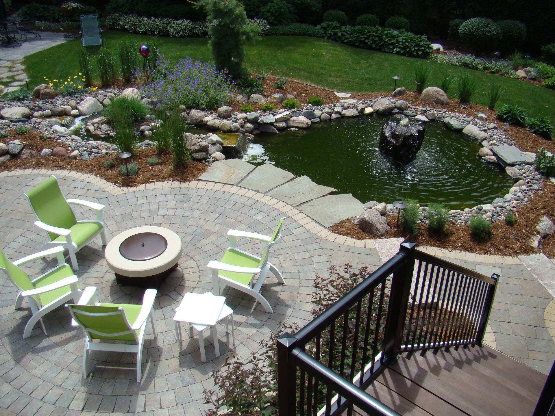 Patio with green chairs around a fire pit, near a pond with fountain, and lush garden.