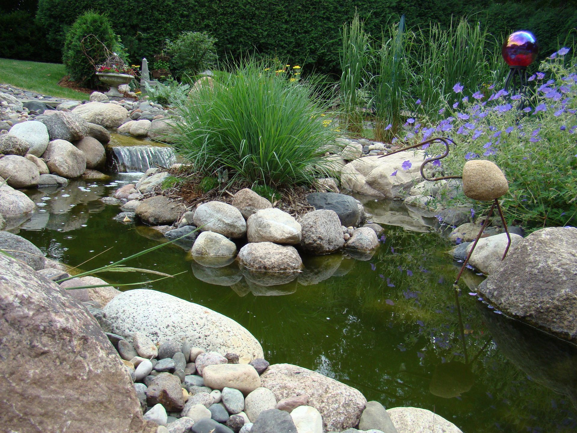 A small pond with rocks, plants, and a waterfall. Green water, bushes, and a sculpture are also visible.