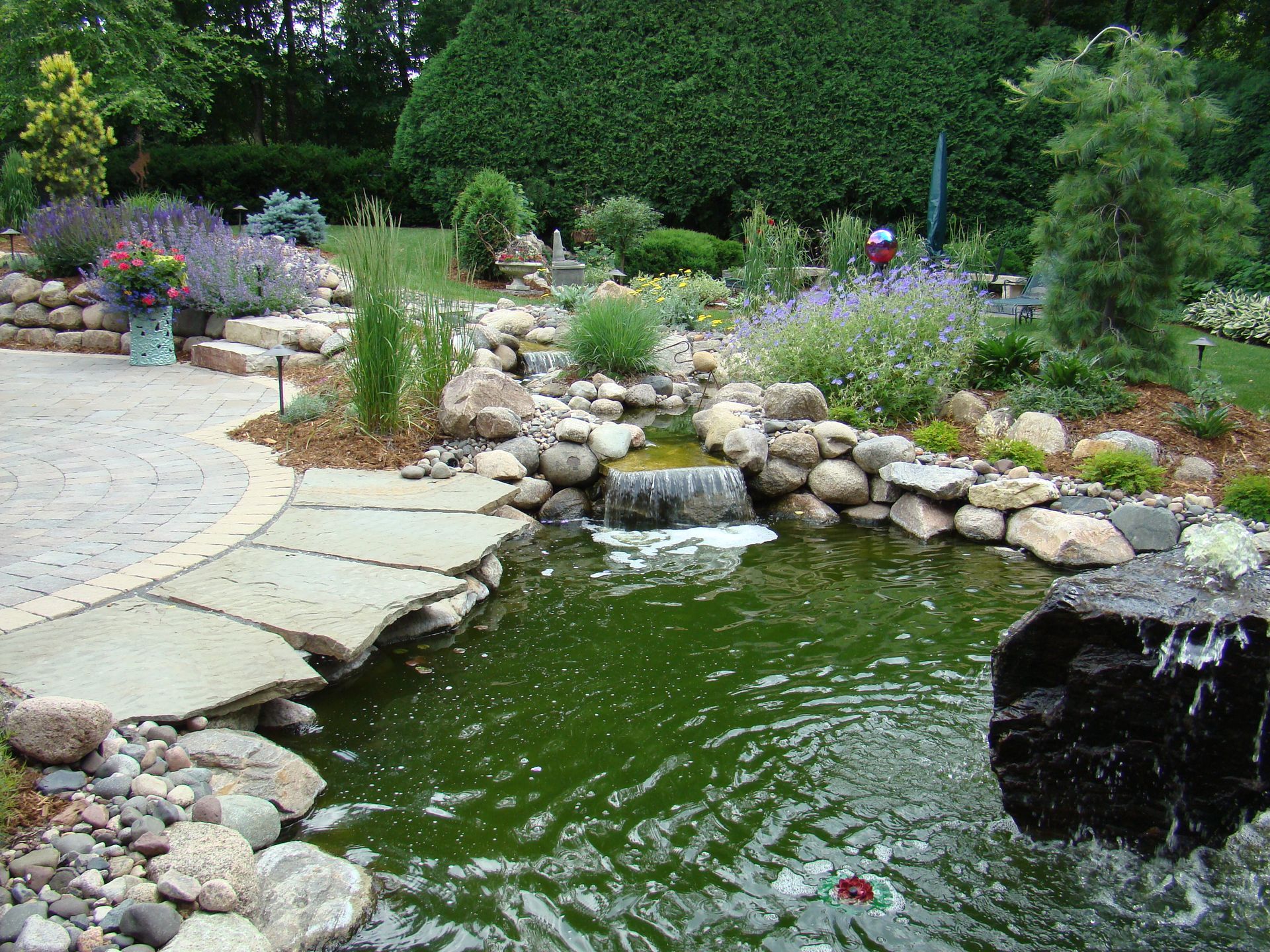 Stone garden pond with waterfall, stepping stones, and surrounding flowers.