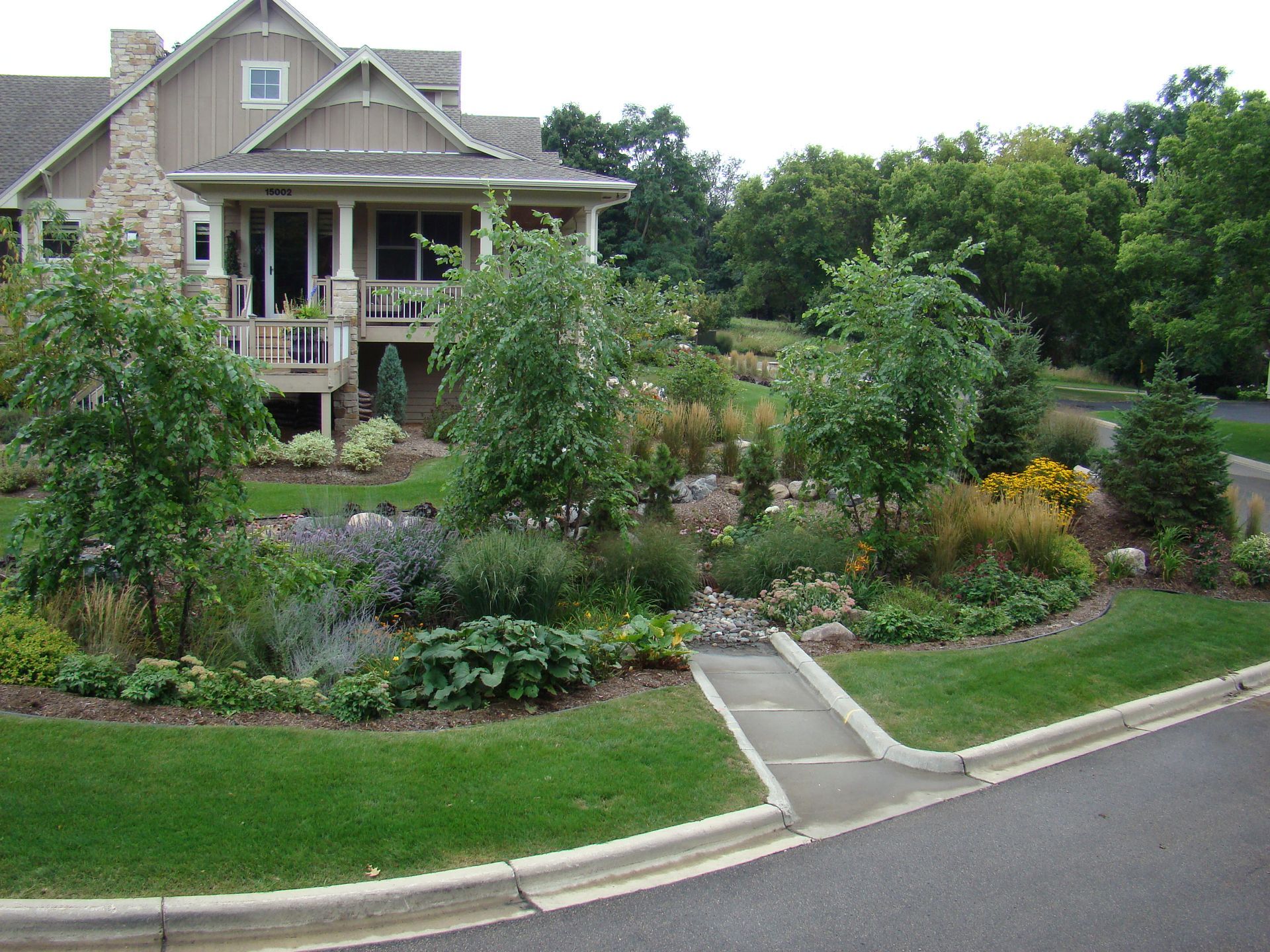 House with lush garden, various trees, and stone accents. Curved landscaping next to road.