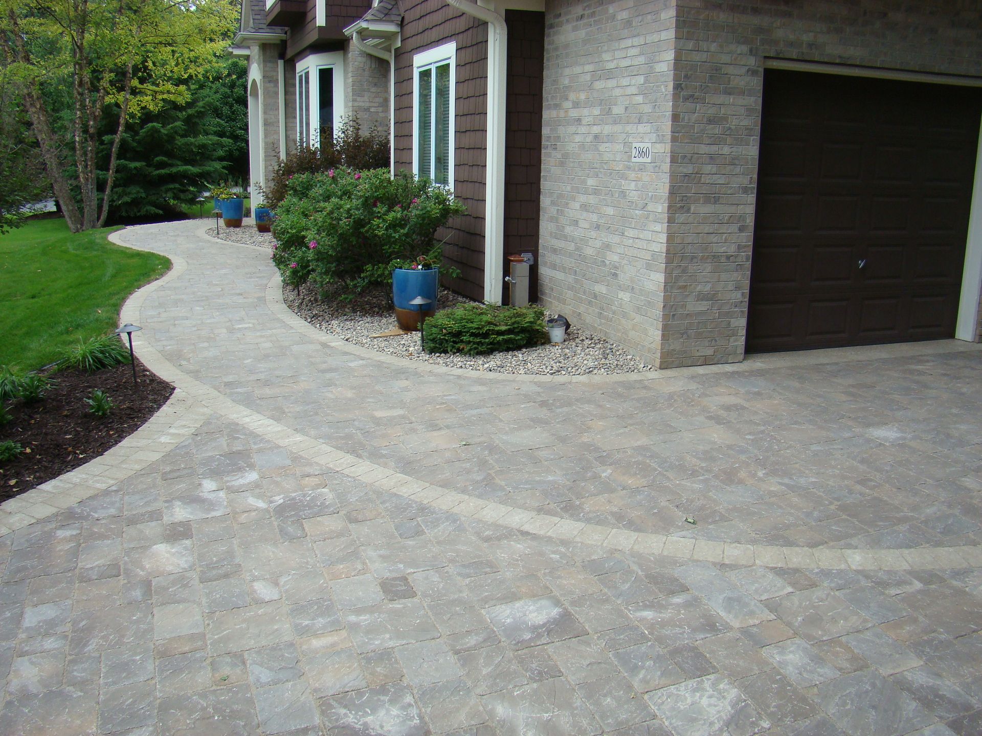 Paver walkway curves past a house with a garage. Brown garage door, tan brick facade, and green landscaping.