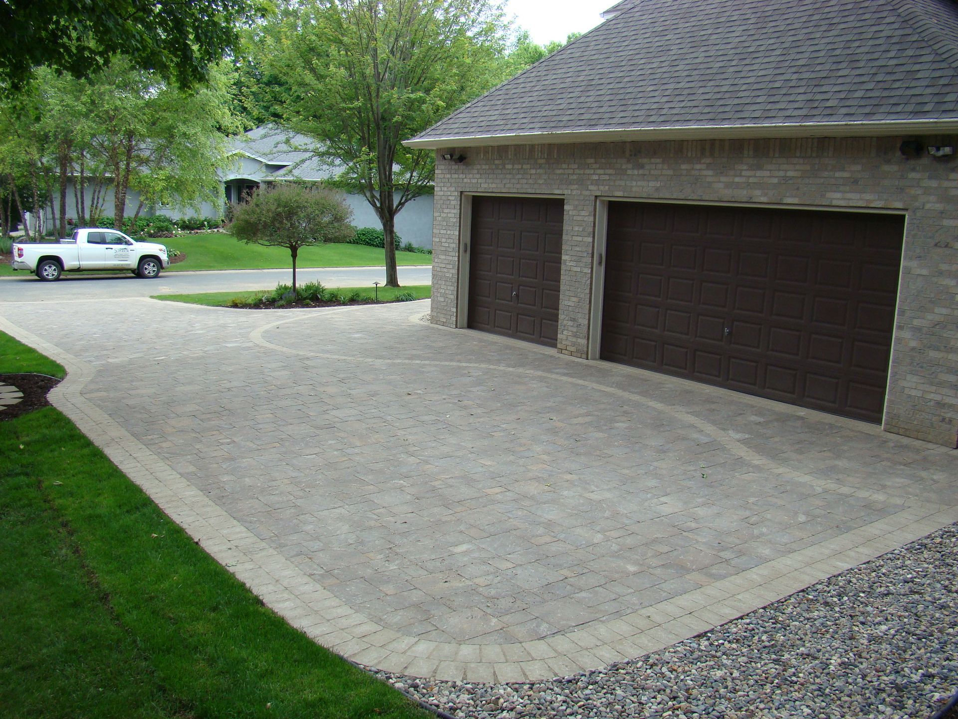 Brick driveway leading to a two-car garage with brown doors, gravel border, and a white pickup truck on the road.