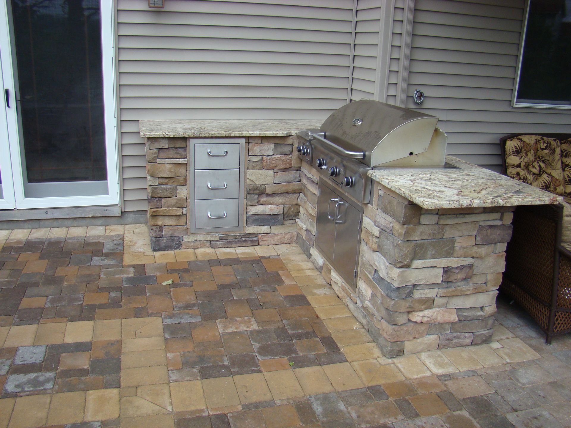 Outdoor kitchen with grill and stone countertop on a brick patio.