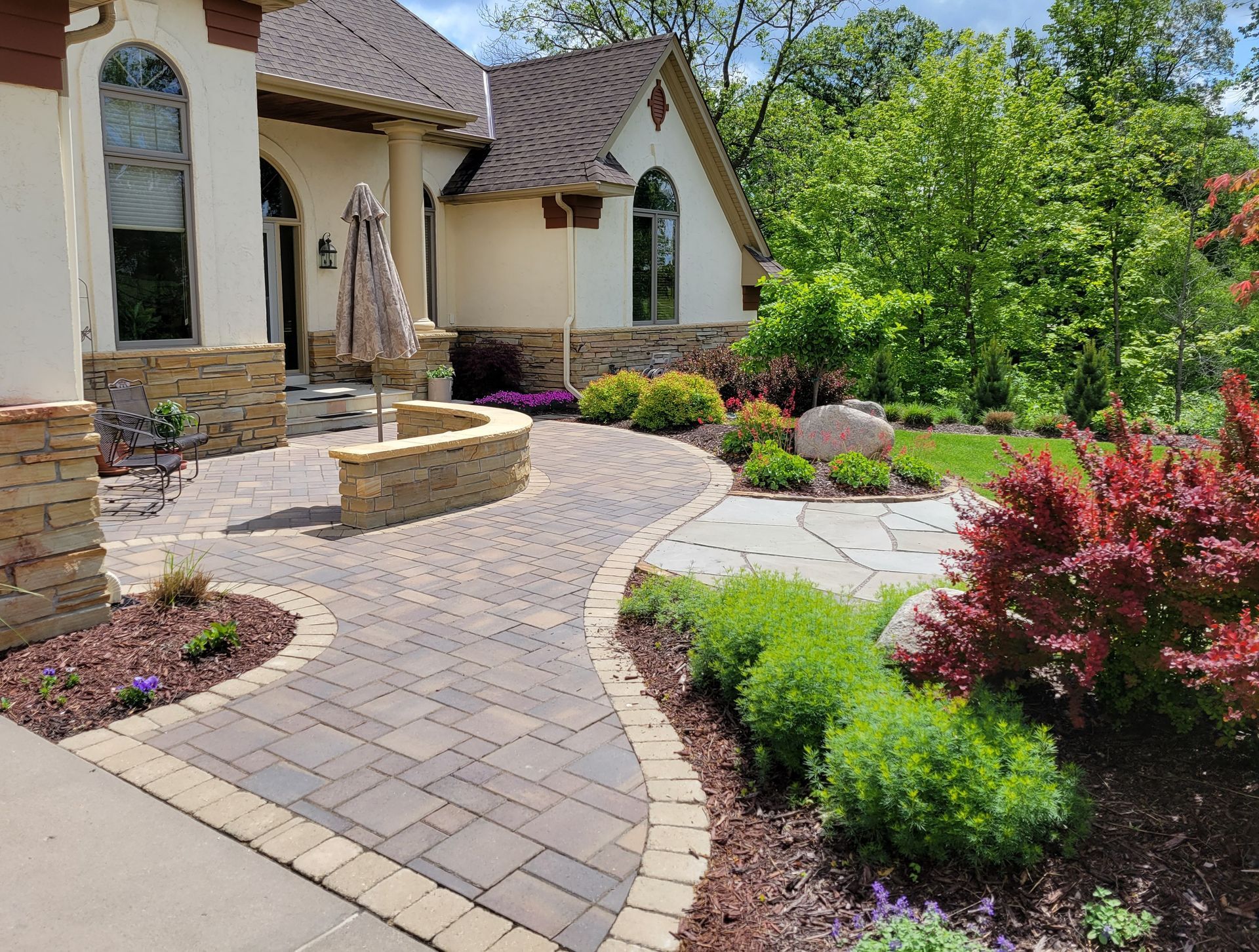 Stone pathway curves through landscaped yard with house in the background; lush greenery and colorful bushes surround the path.