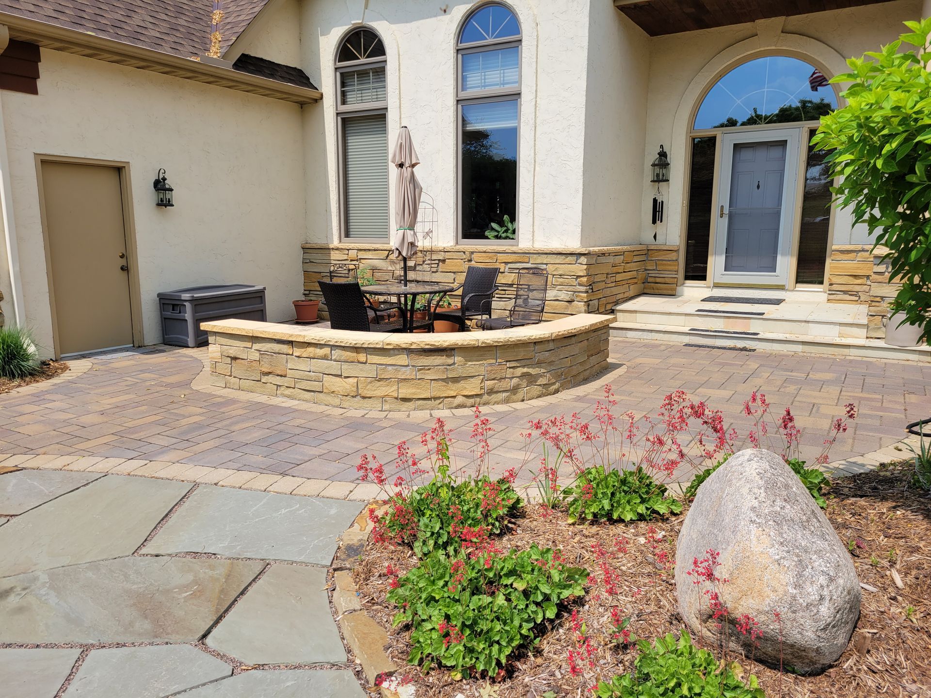 Stone patio and landscaping in front of a tan stucco house with an arched entry.