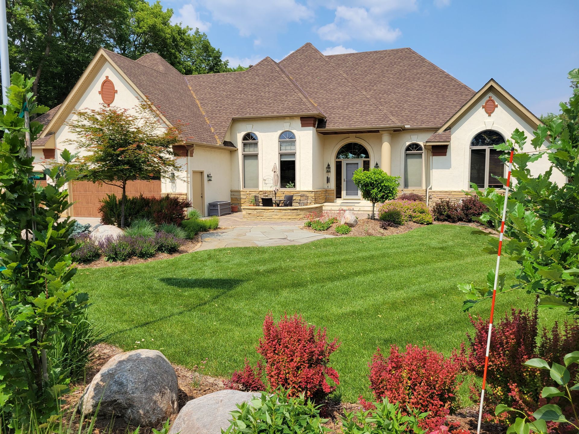 Beige house with brown roof and green lawn, surrounded by landscaping on a sunny day.