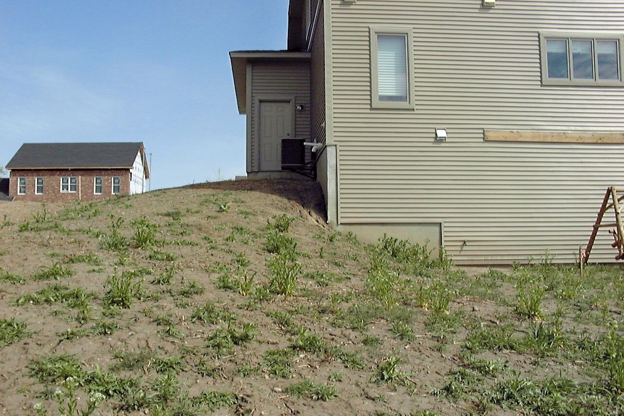Hill of dirt and weeds in front of a light-colored building with a dark-roofed building in the background.