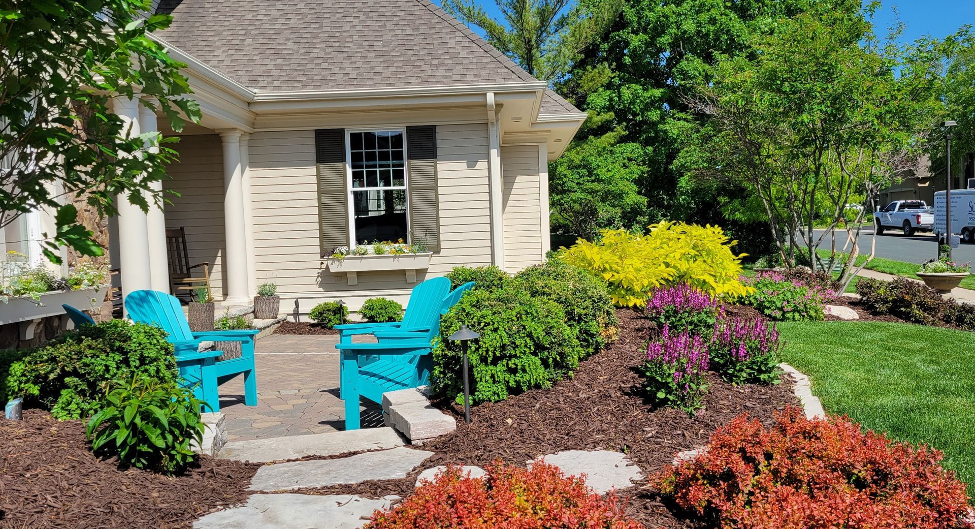 Two turquoise Adirondack chairs on a stone path in front of a house, surrounded by colorful flowers and landscaping.