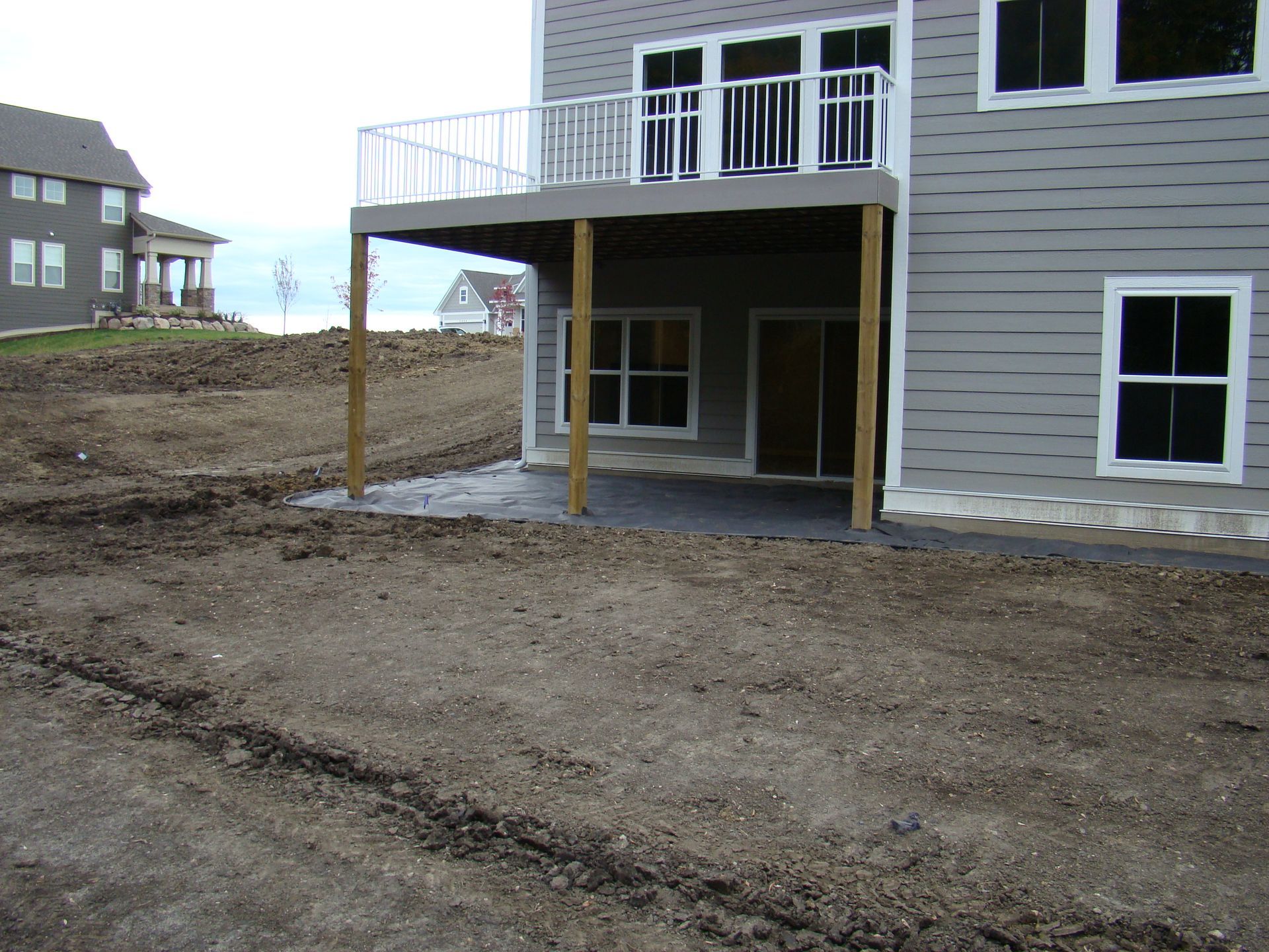 Back of a new house with a deck above a concrete patio, surrounded by freshly graded dirt.