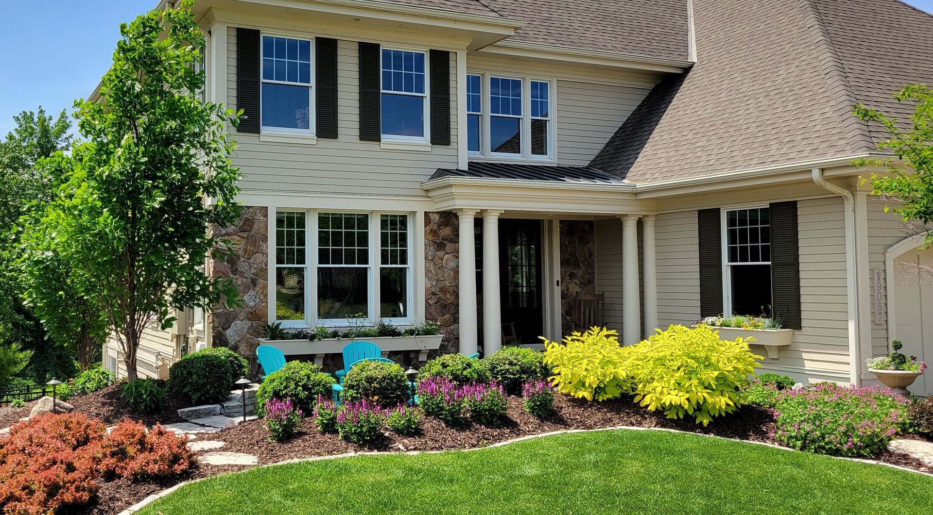 Two-story house with green lawn, flower beds, and blue chairs. Light siding, dark shutters, and a sunny day.