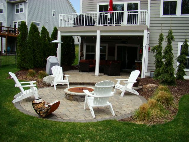 Backyard patio with fire pit, white chairs, and a two-story house with a deck above.