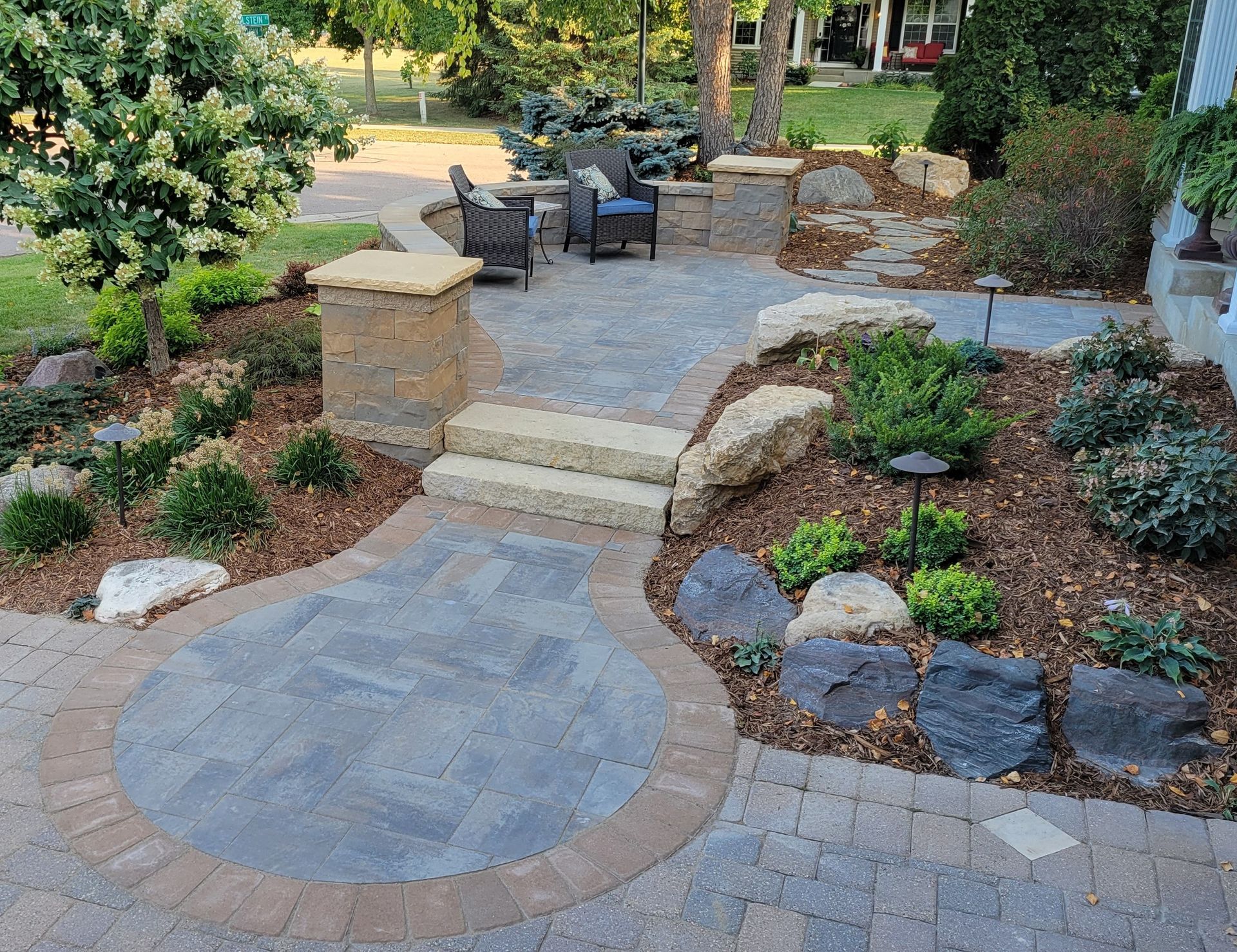 Stone patio with seating area, steps, and landscaping. Brown and gray pavers, green shrubs, mulch.