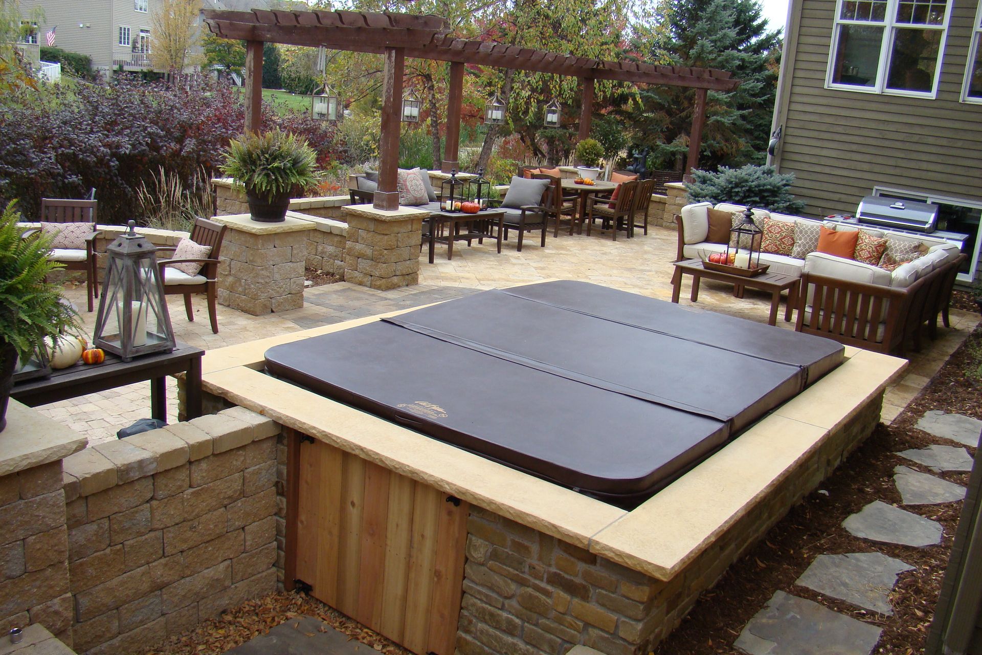 Backyard patio with hot tub, seating areas, pergola, and decorative stonework. Autumn colors.