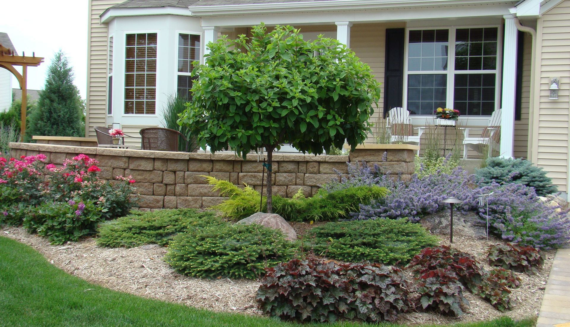 Landscaped yard with retaining wall, small tree, various plants, and beige gravel.