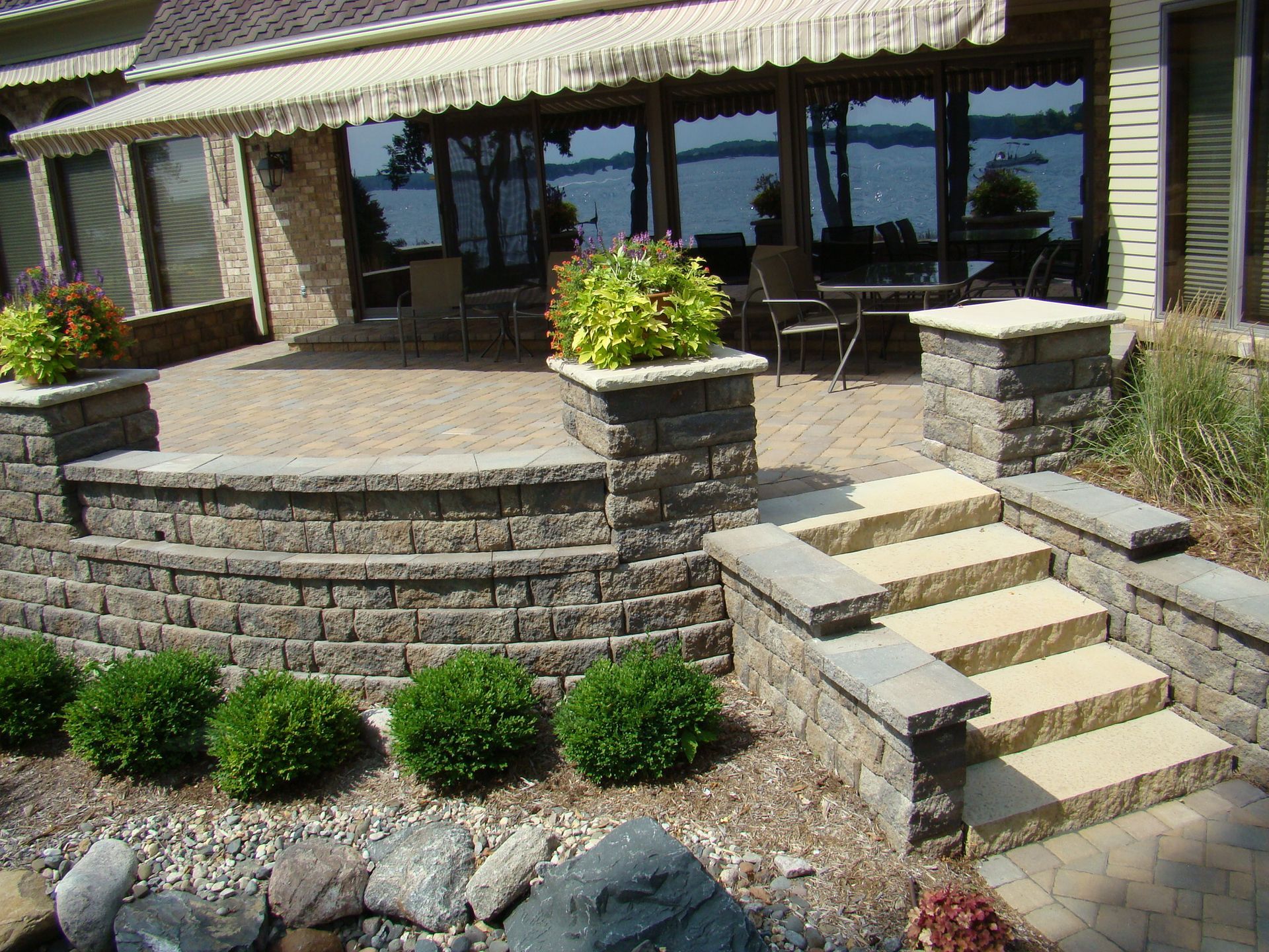 Stone patio with steps, raised planter beds, awning, and lake view.