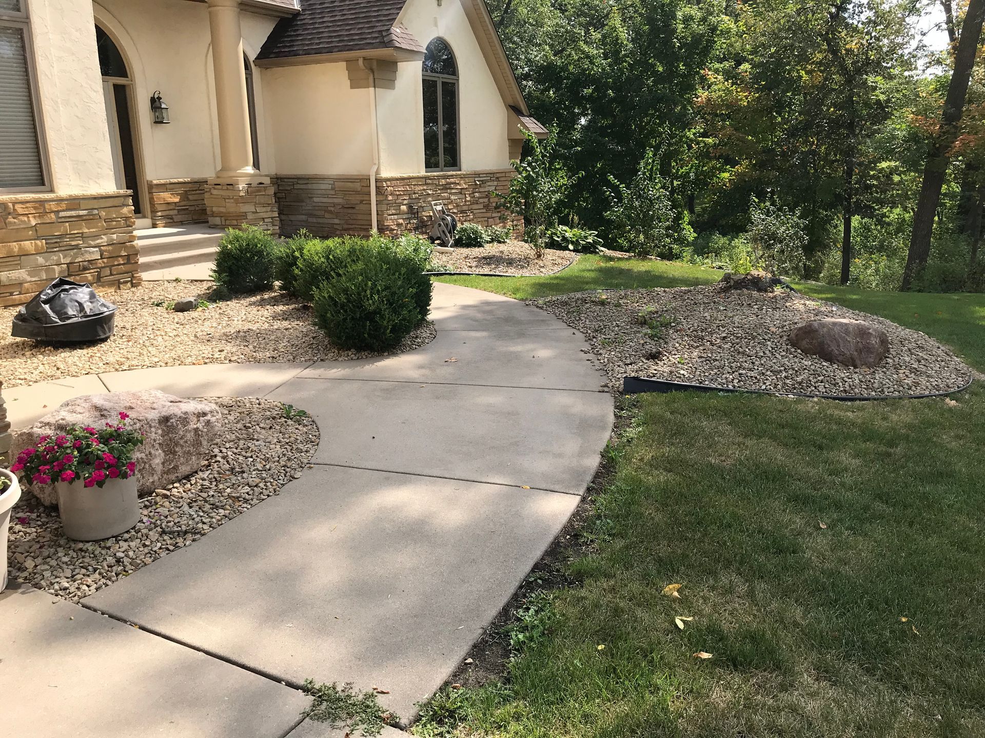 Concrete walkway curving towards a beige house with gravel landscaping, grass, and trees.