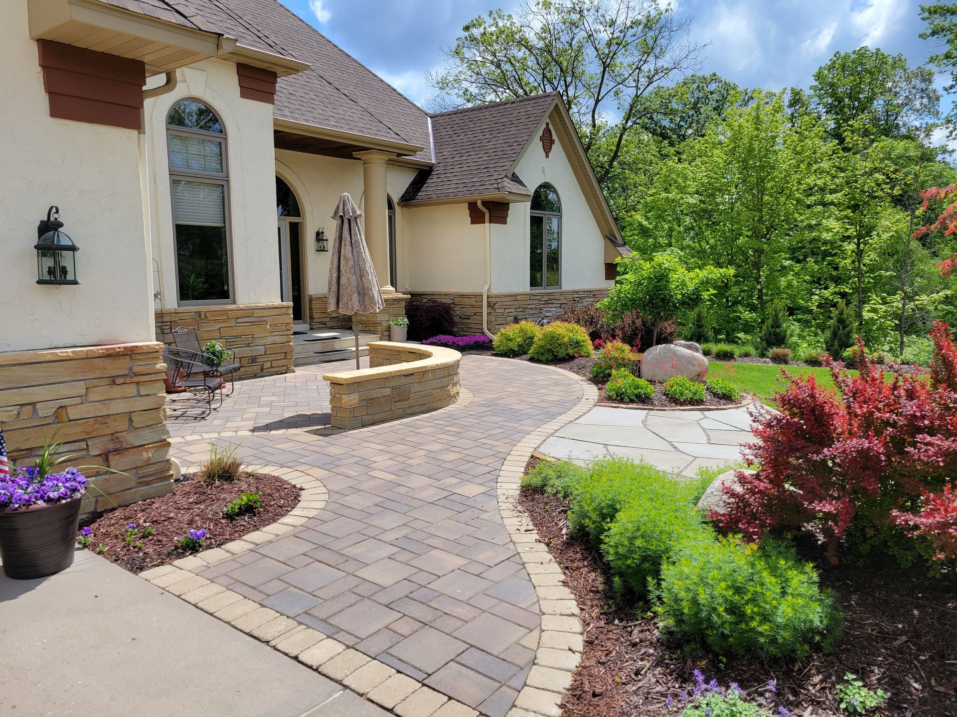 Stone-paved walkway leads to a beige house with arched windows and landscaping, including colorful bushes and trees, on a sunny day.