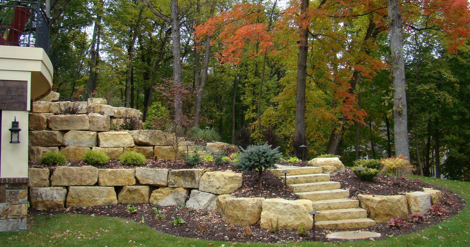 Stone steps and retaining wall in a landscaped yard with autumn foliage in the background.