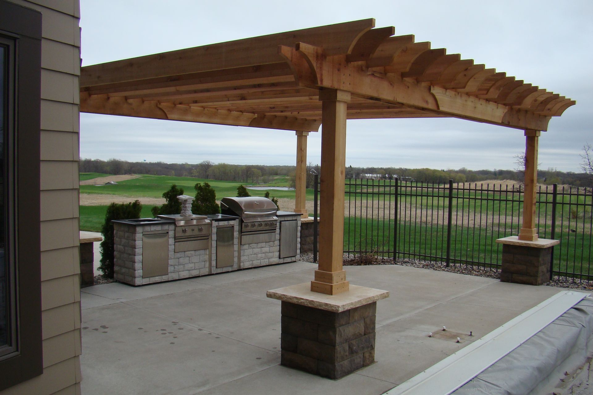 Pergola over outdoor kitchen on a concrete patio overlooking a field; light brown wood structure.
