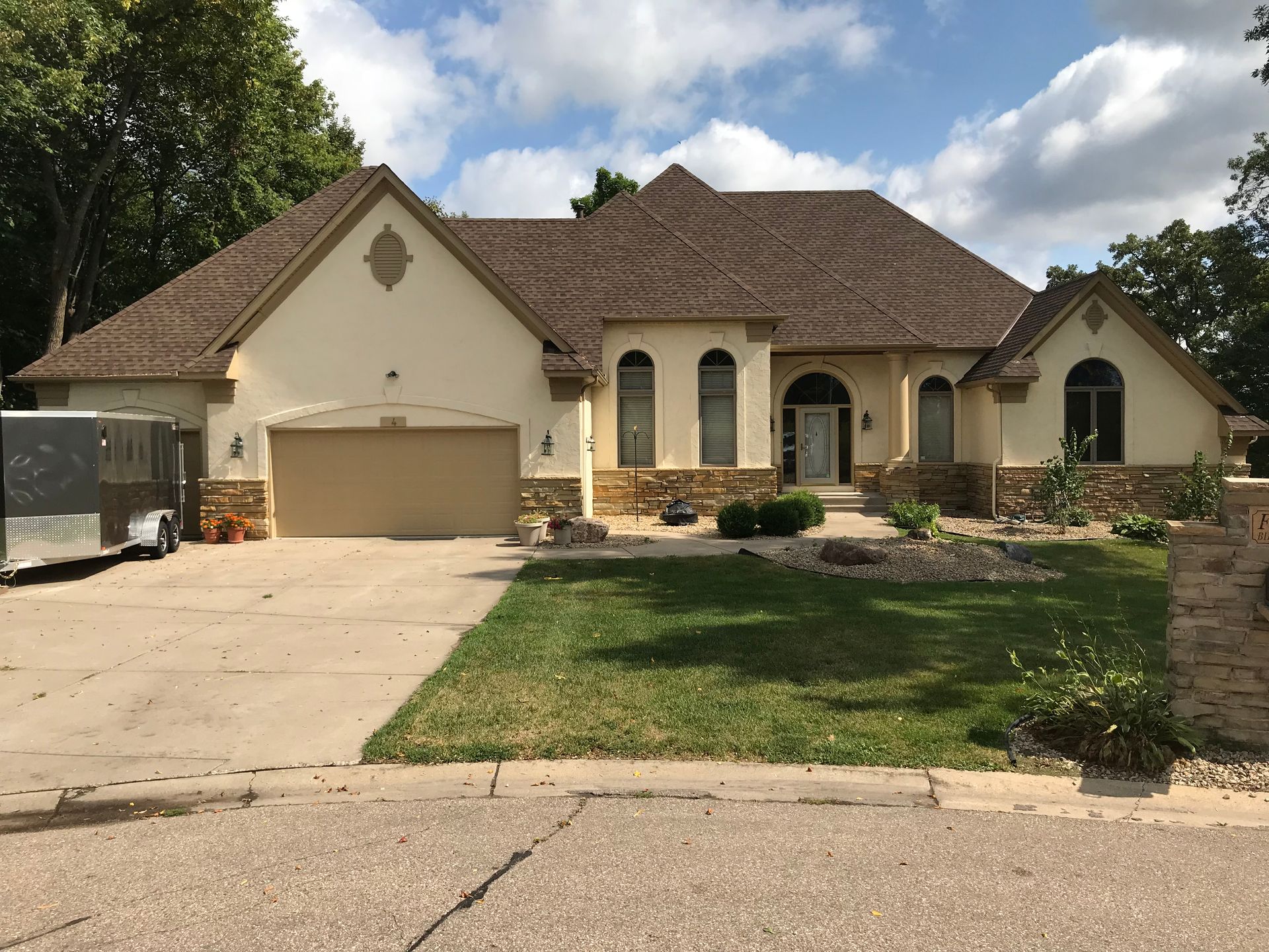 Beige stucco house with brown roof and garage, stone accents, and green lawn. Trailer parked in driveway.