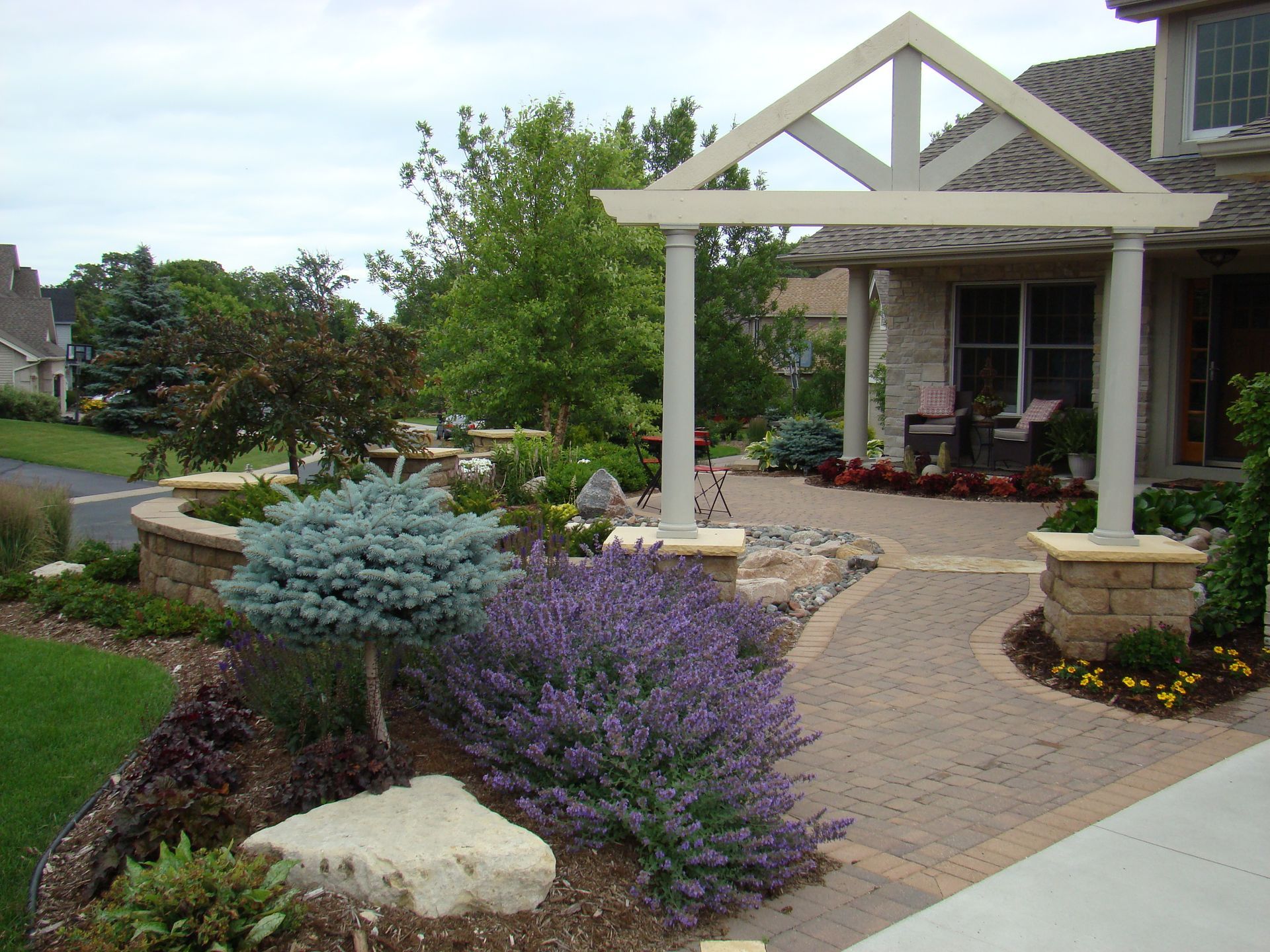 Stone pathway leading to a house with a pergola-covered entrance, surrounded by colorful landscaping.