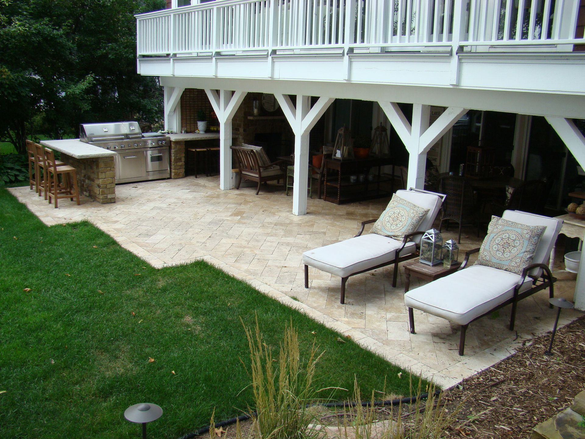 Outdoor patio with lounge chairs under a white deck. Barbecue and bar area under the deck, next to green grass.