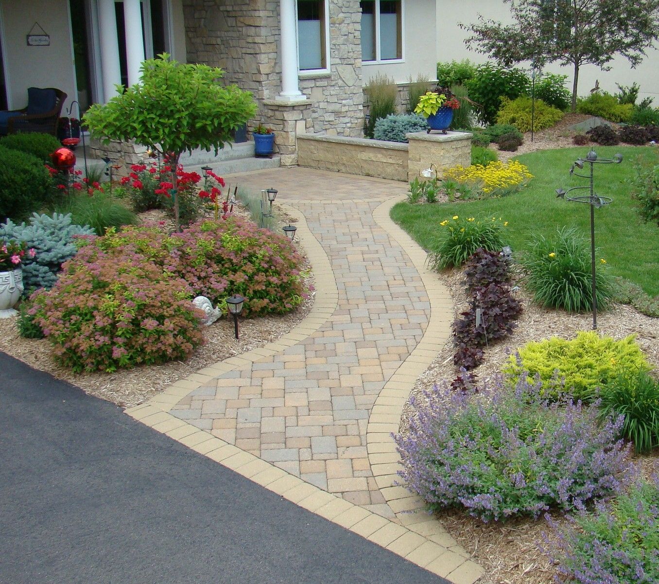 Brick walkway curves through a landscaped front yard with colorful flowers, shrubs, and trees.