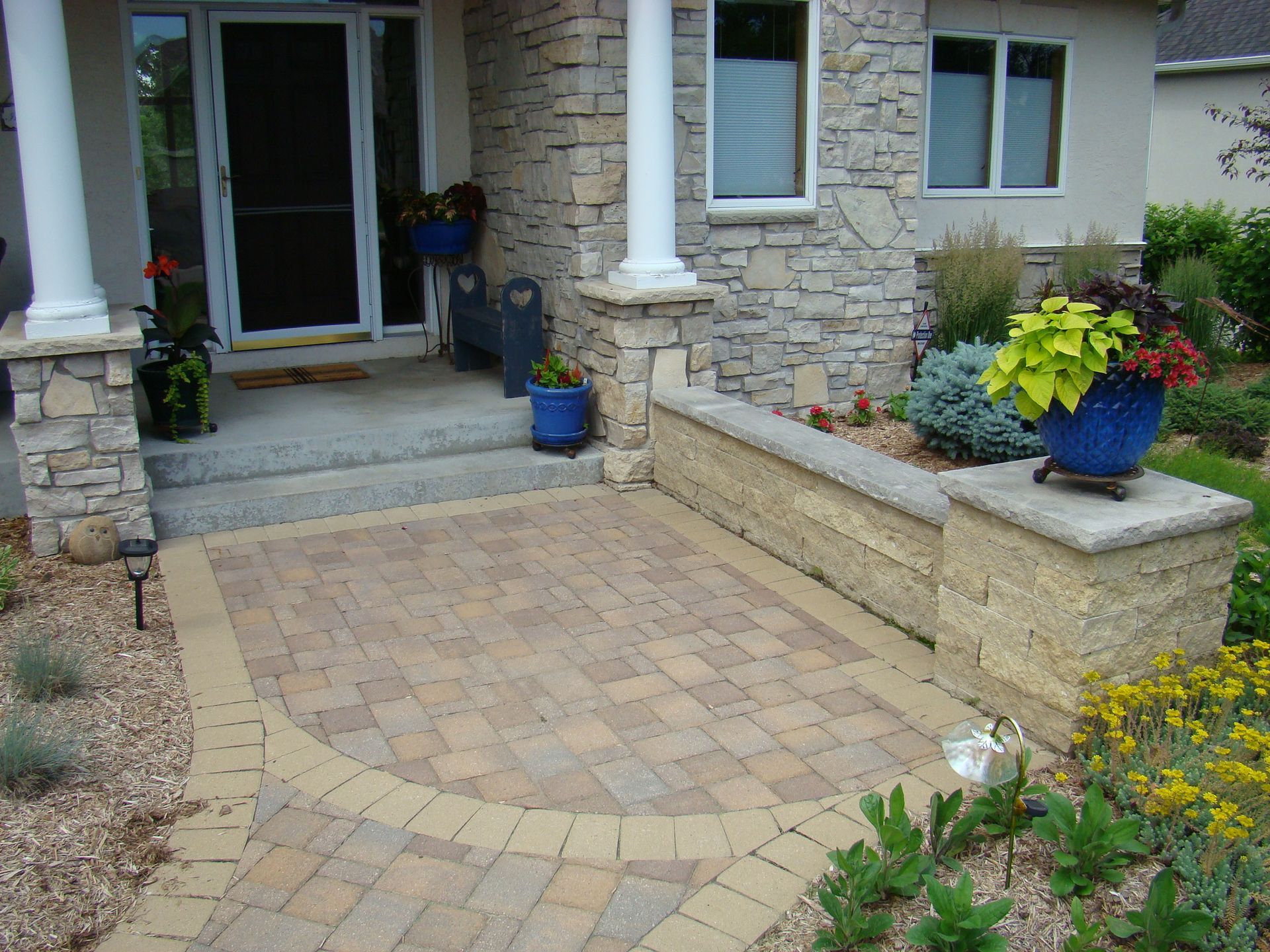 Brick patio with a curved edge, leading to a home's entryway with planters and stone facade.