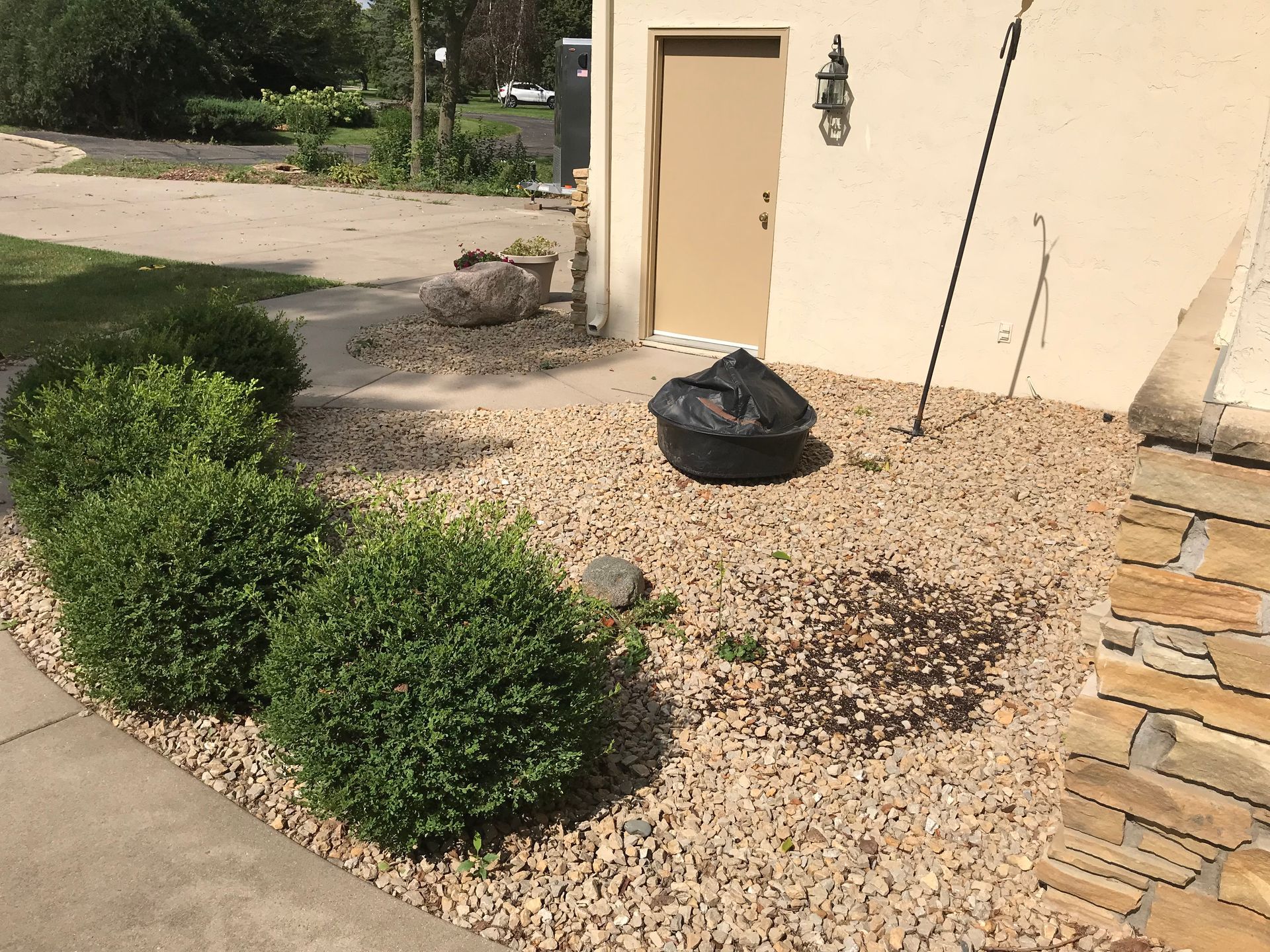 Bushes in a gravel bed beside a building with a tan door. Black trash bag is also in the gravel.