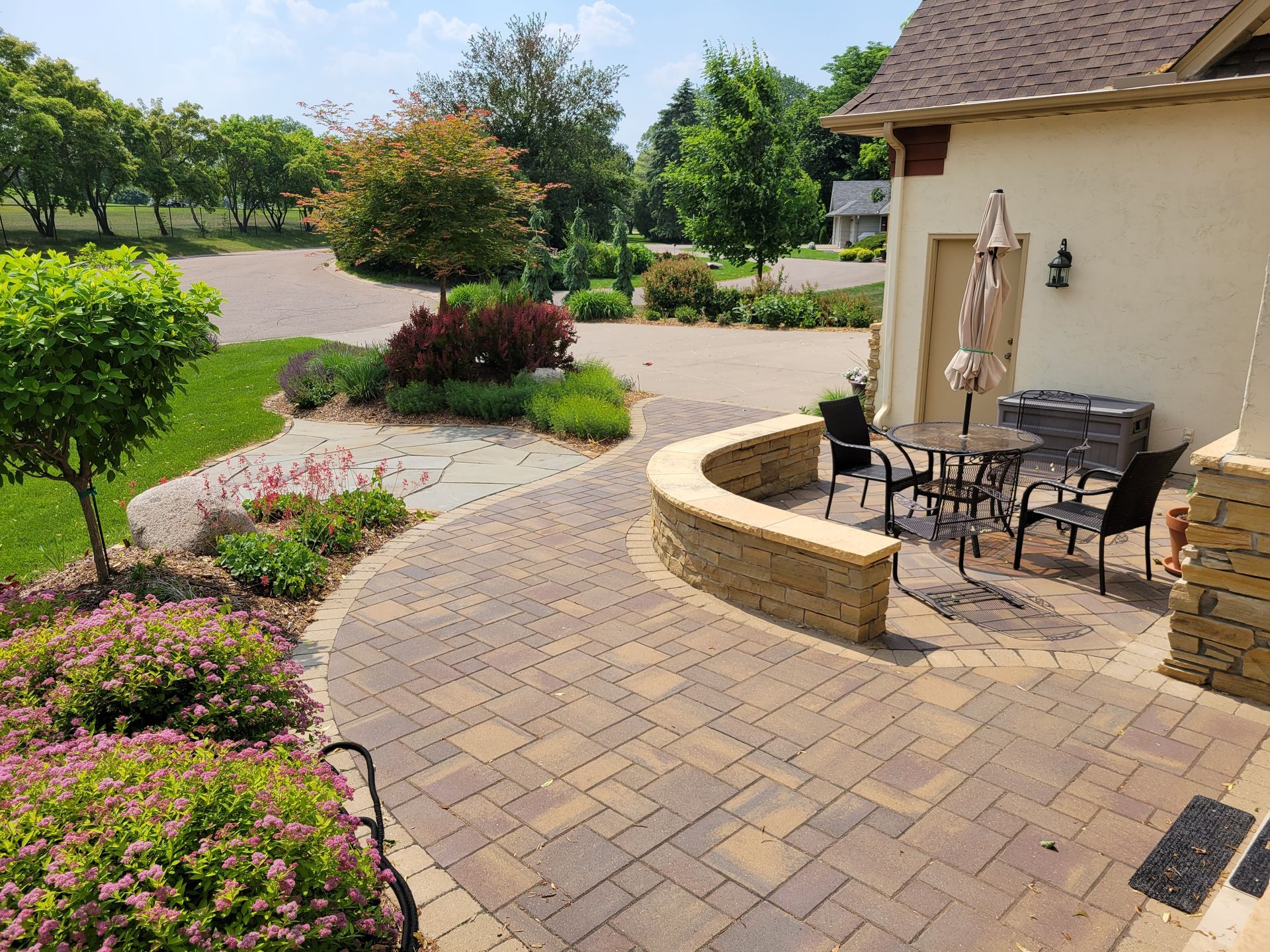 Brick patio with a curved stone wall, table, chairs, and garden with trees. Sunny day.