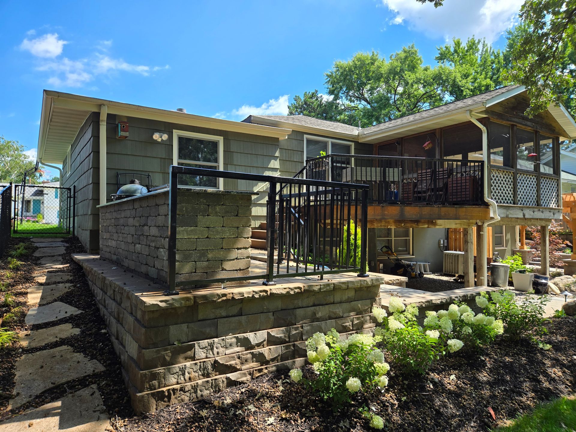 Backyard with a gray house, retaining wall, black railing, and a screened porch under a blue sky.