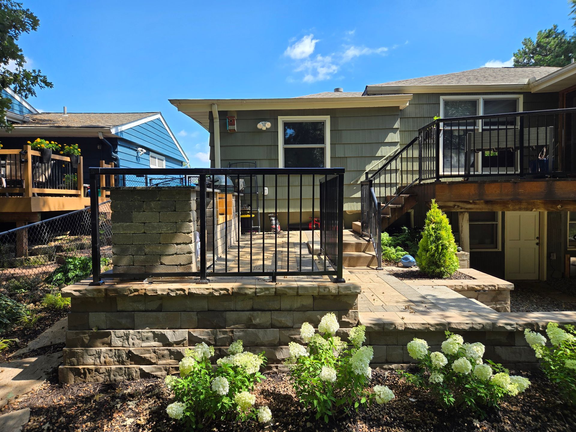 Backyard patio with black railing, stone wall, and landscaping under a blue sky.