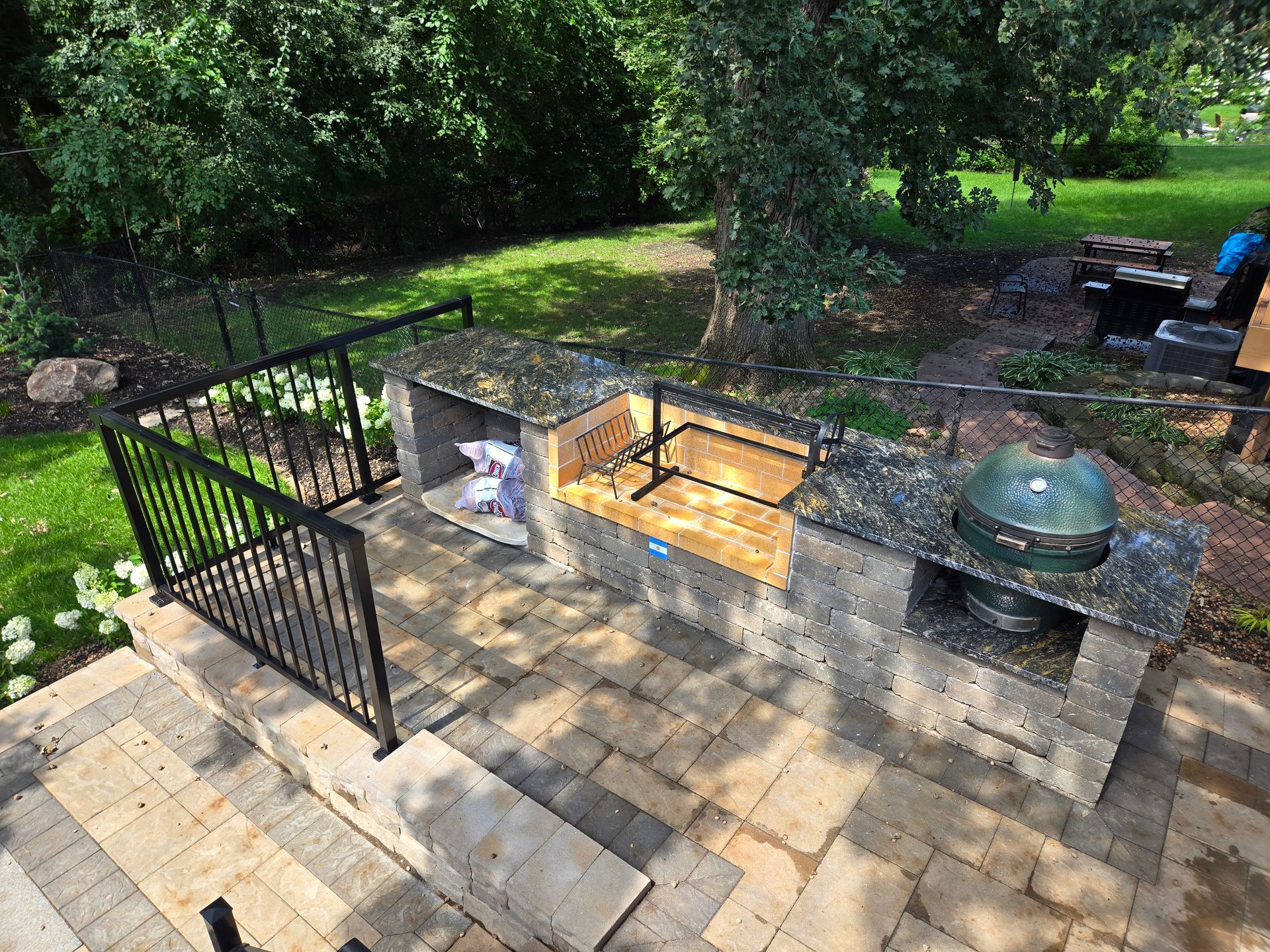 Outdoor kitchen with built-in grill, smoker, and countertops on a stone patio. Black metal railing on stairs.
