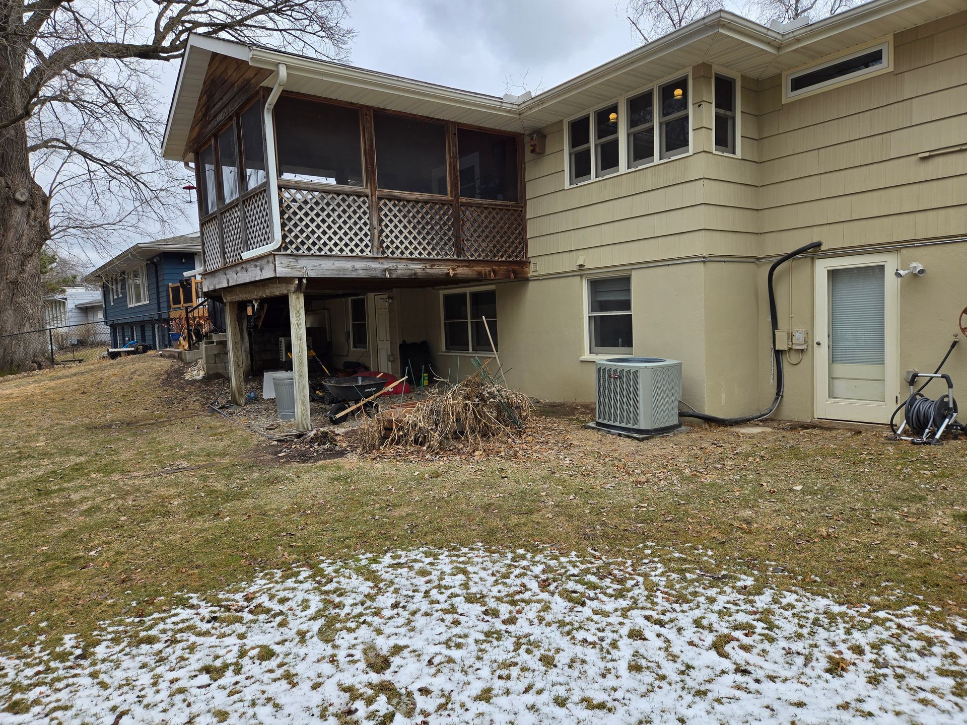 Backyard view of a two-story beige house with a screened porch, grass, some snow, and other houses.