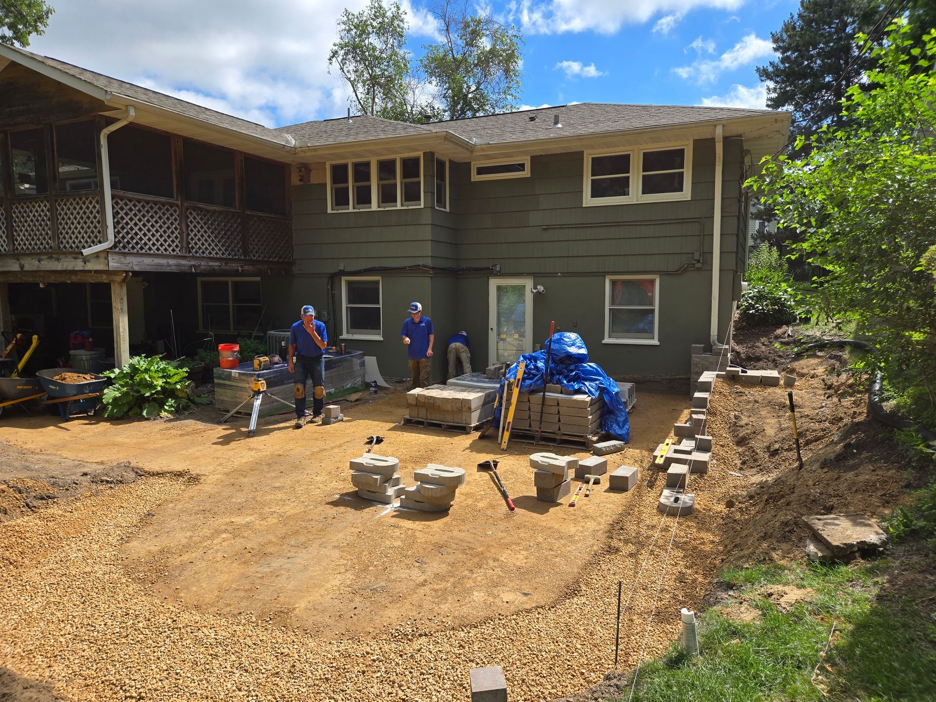 Backyard construction site: workers laying pavers near a two-story green house with a screened porch on a sunny day.