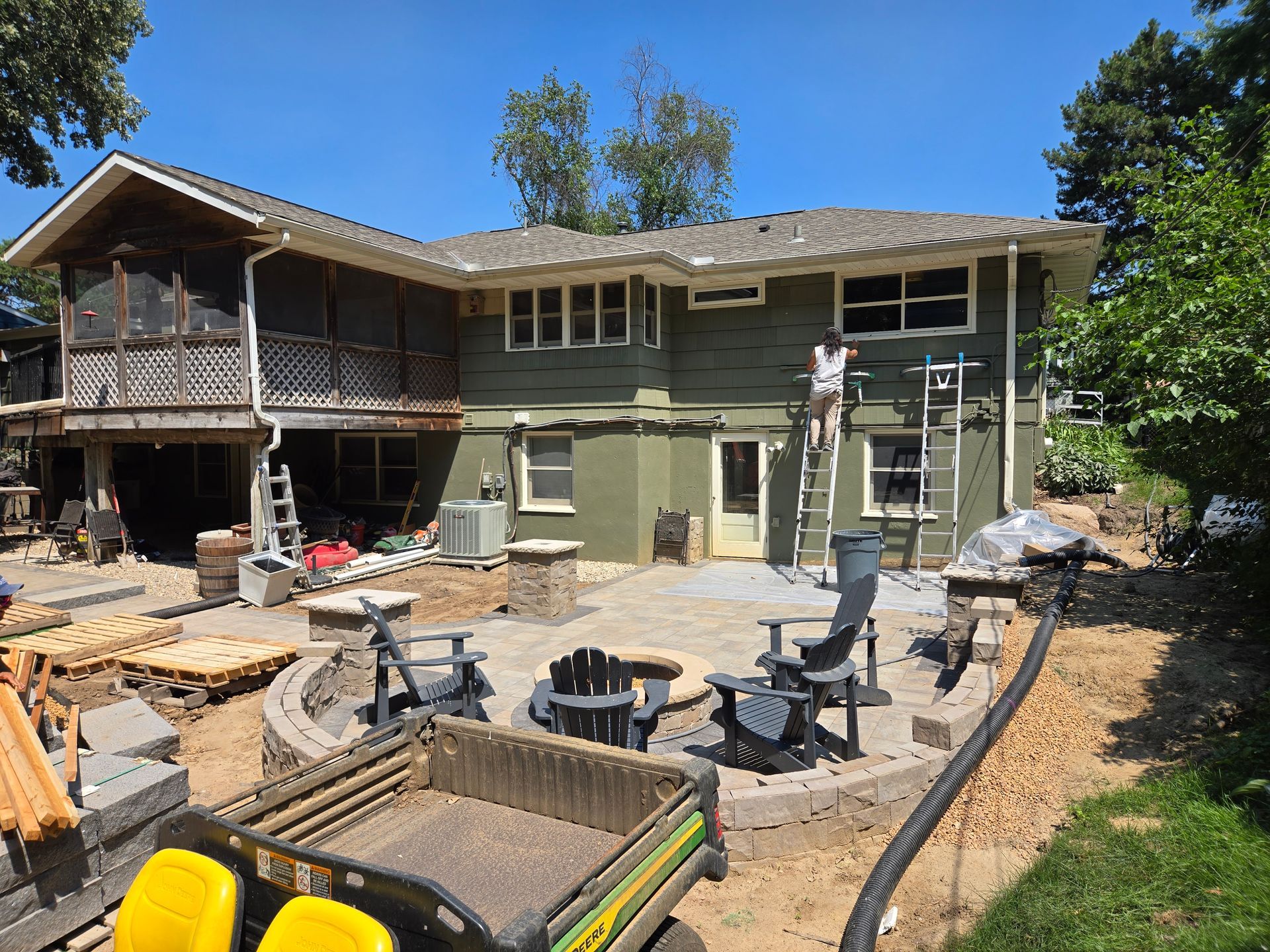 Backyard renovation: Green house with patio, fire pit, and a person on a ladder, construction materials visible.