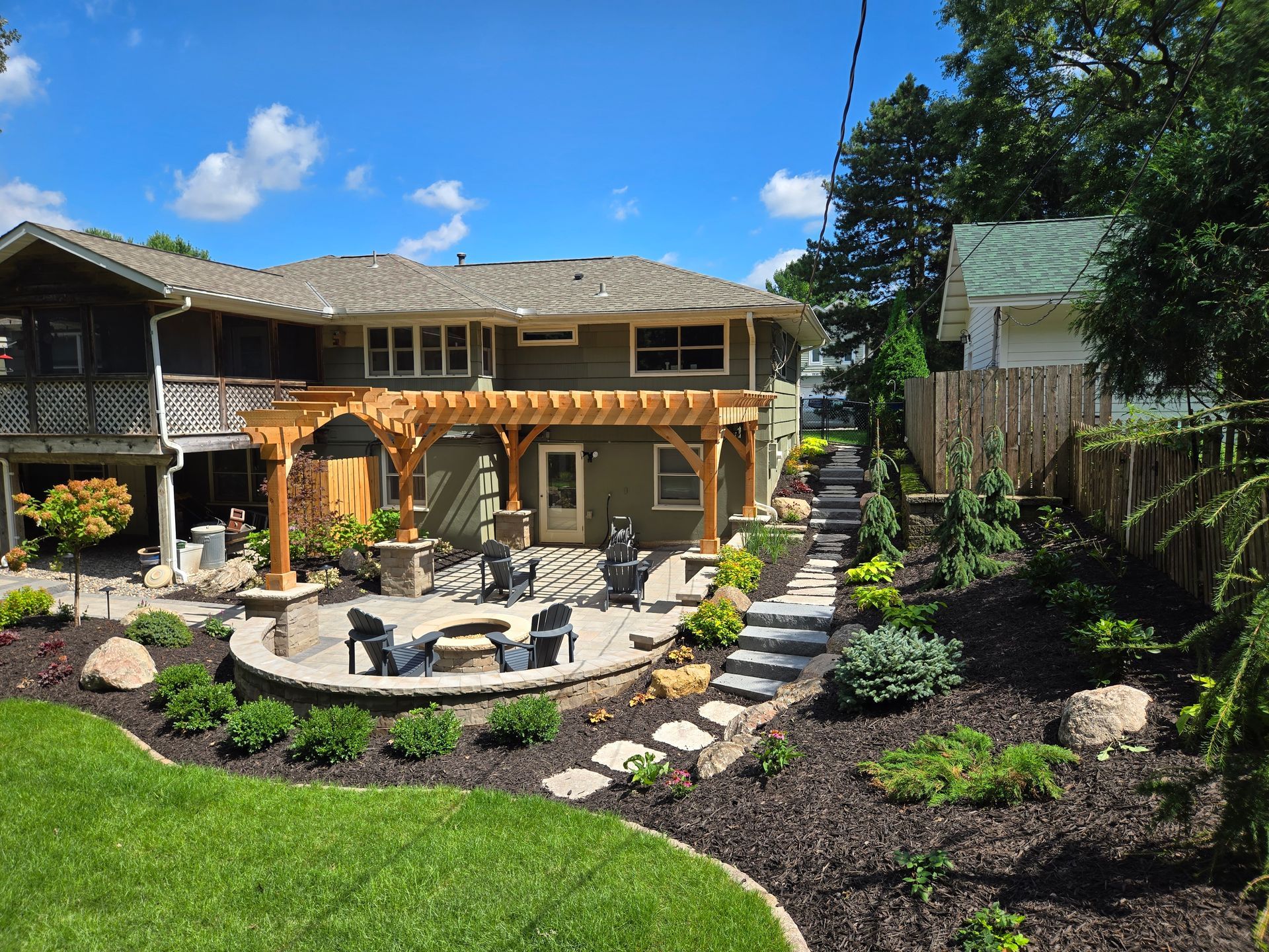 Backyard patio with pergola, stone fire pit, seating, steps, and landscaping near a two-story house.