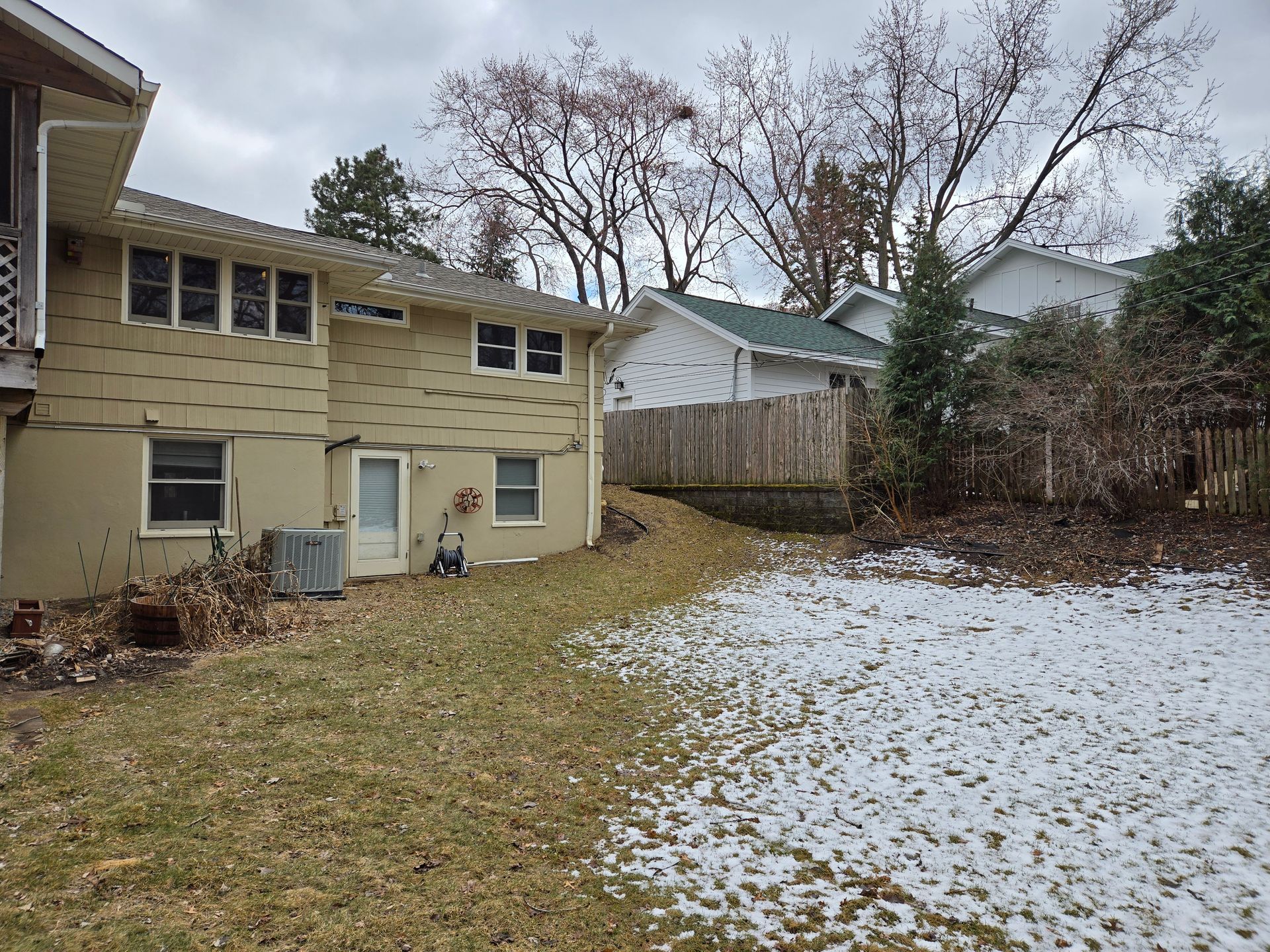 Backyard view of a two-story beige house and a snowy lawn on a cloudy day.