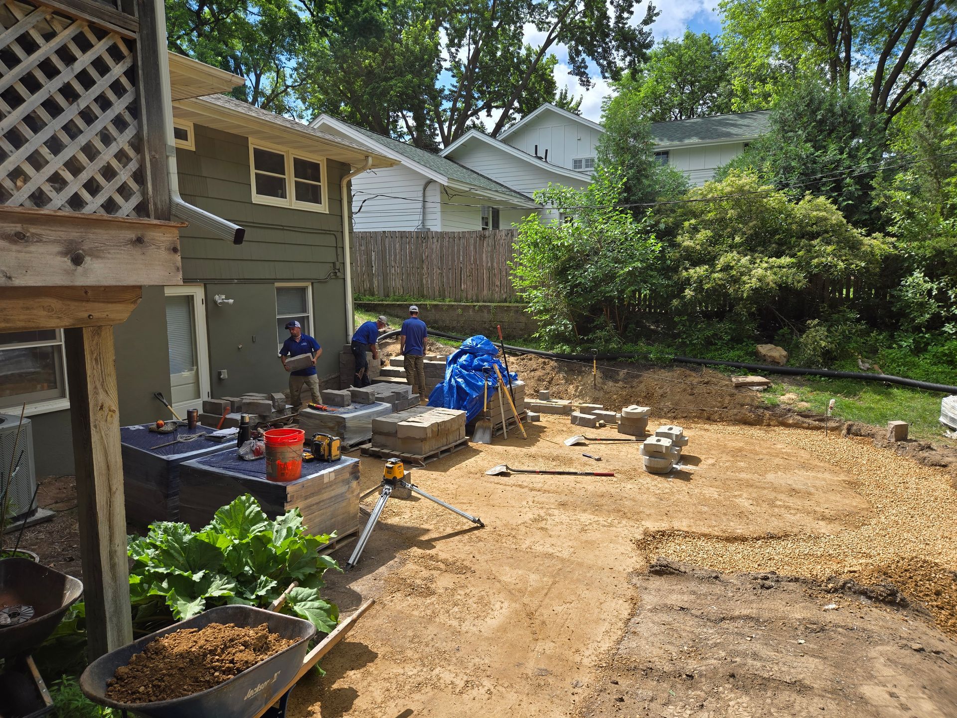 Workers constructing a patio in a backyard. Bricks, tools, and dirt surround the house.