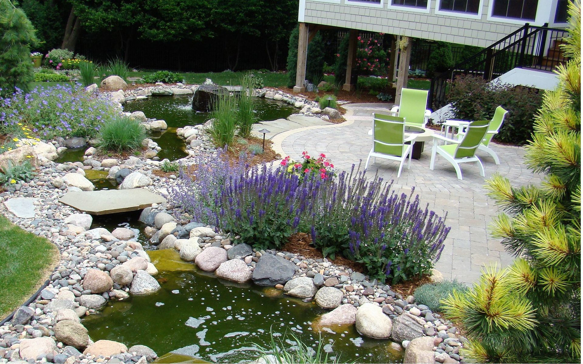Landscaped backyard with ponds, stone path, green chairs, and purple flowers.