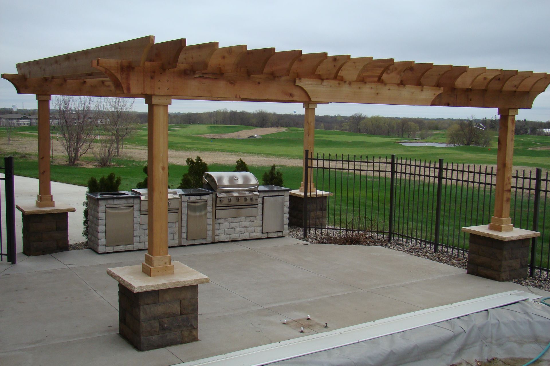 Outdoor kitchen with wooden pergola over grill and counter on concrete patio.