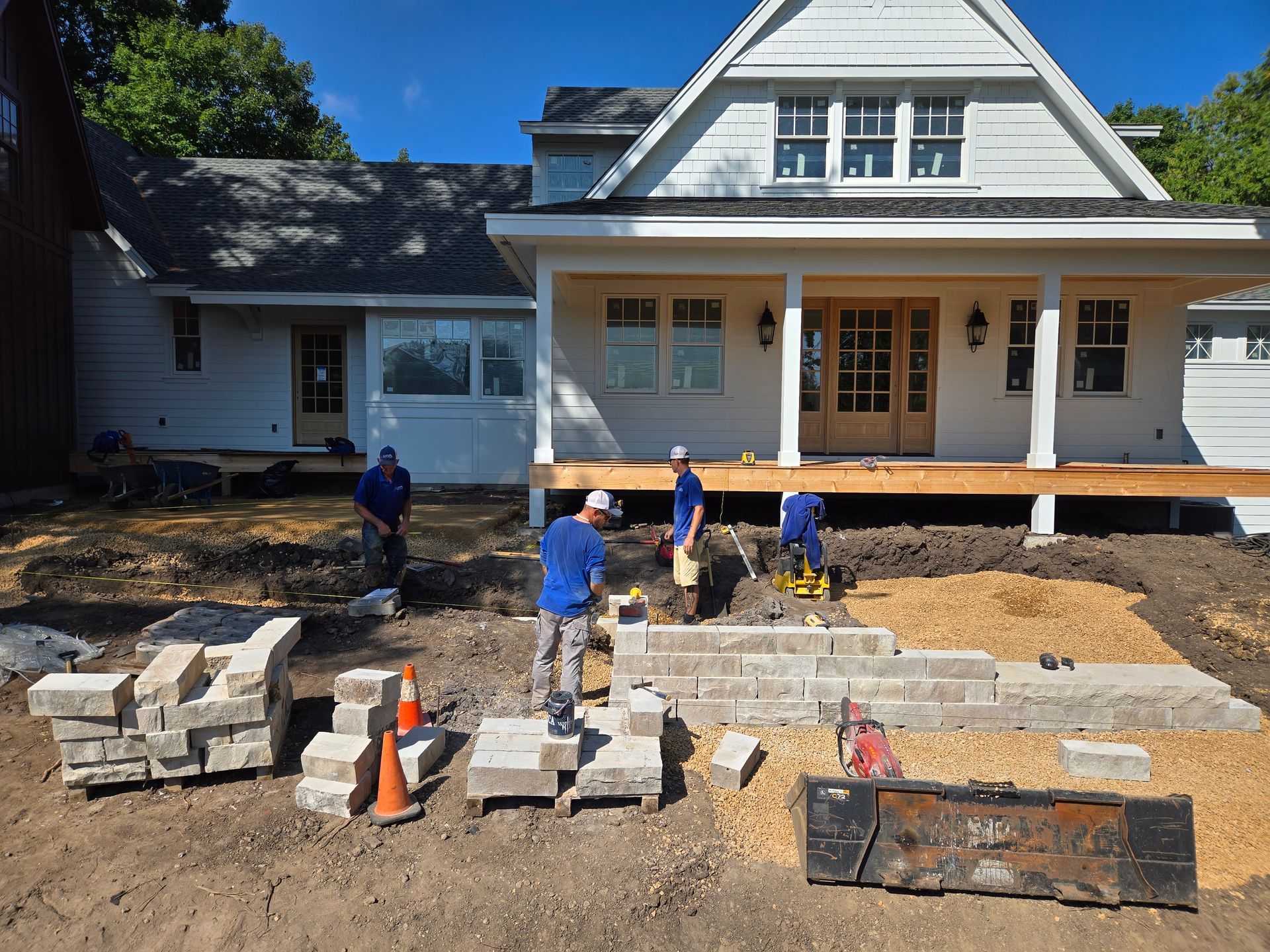 Construction workers building a stone wall in front of a white house with a porch, under a blue sky.