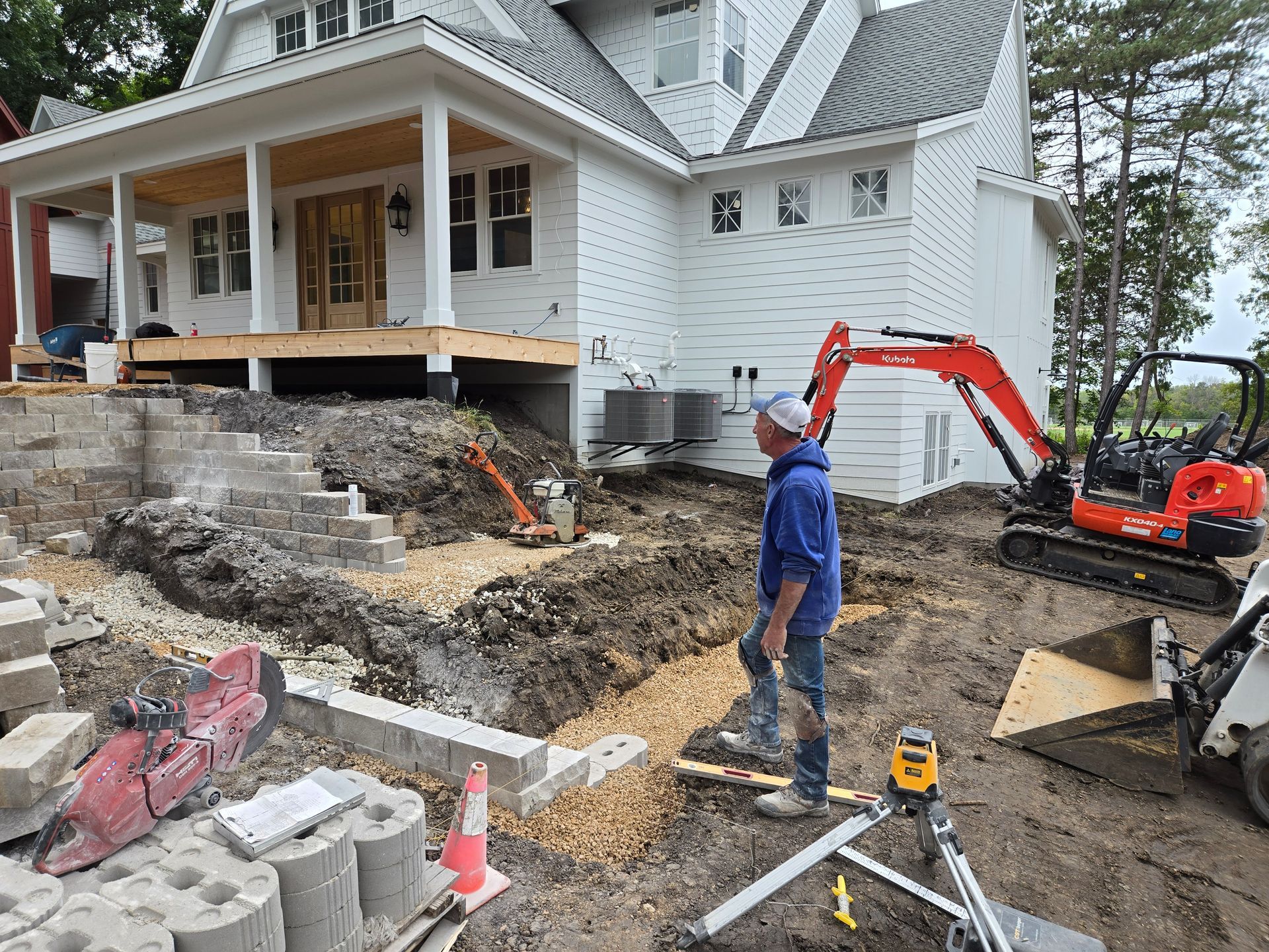 Construction site: man working near a white house with a porch, excavator, and retaining wall being built.