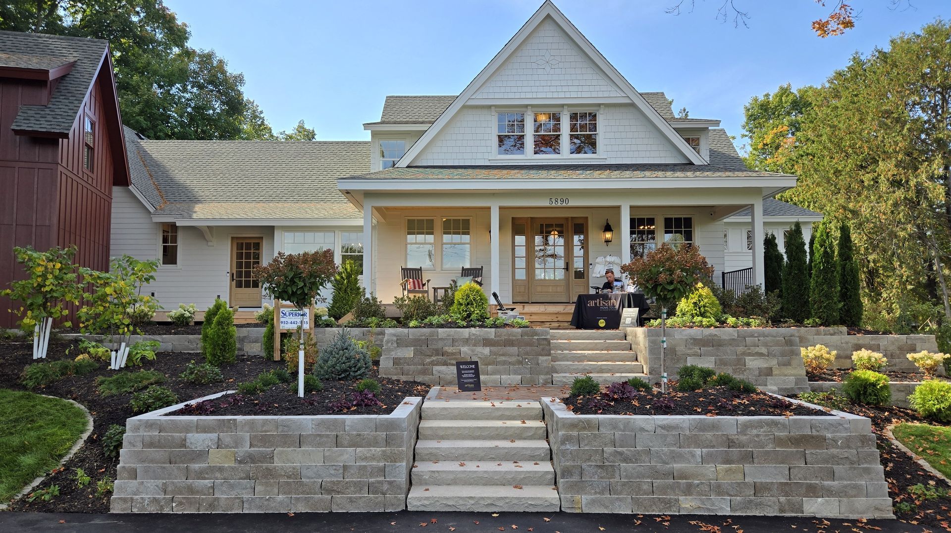 White house with porch, tiered stone walls, and steps leading to the entrance. Lush landscaping with trees and shrubs.