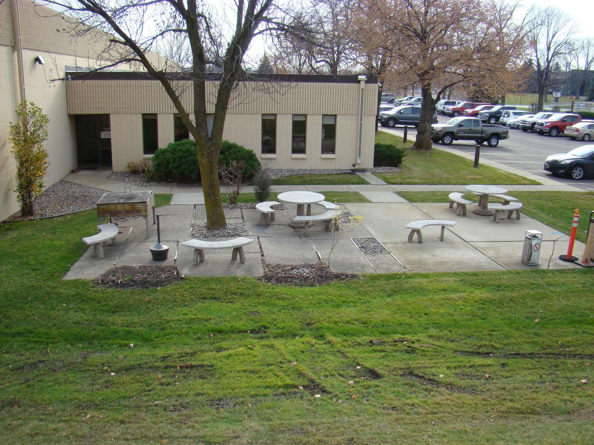 Outdoor concrete picnic tables and benches in front of a tan building; parked cars in the distance.