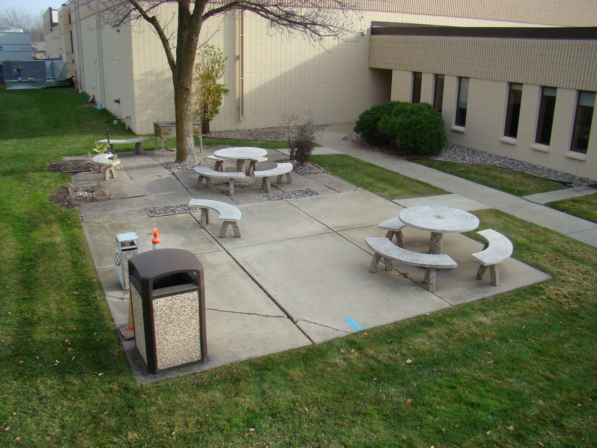 Courtyard with concrete tables, benches, trash can, and patches of grass near a brick building.