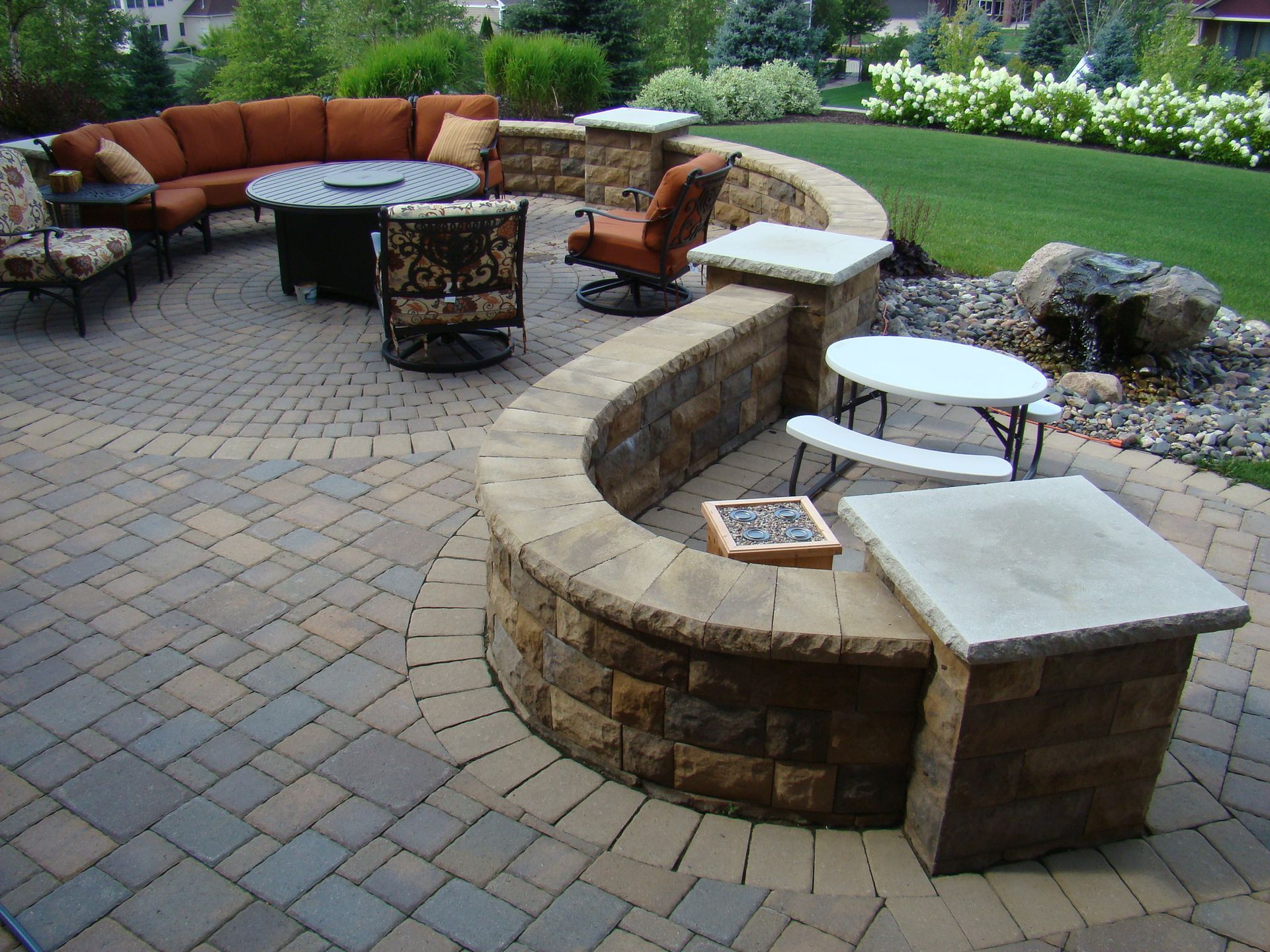 Brick patio with curved retaining wall, outdoor seating area, fire pit, and water feature.